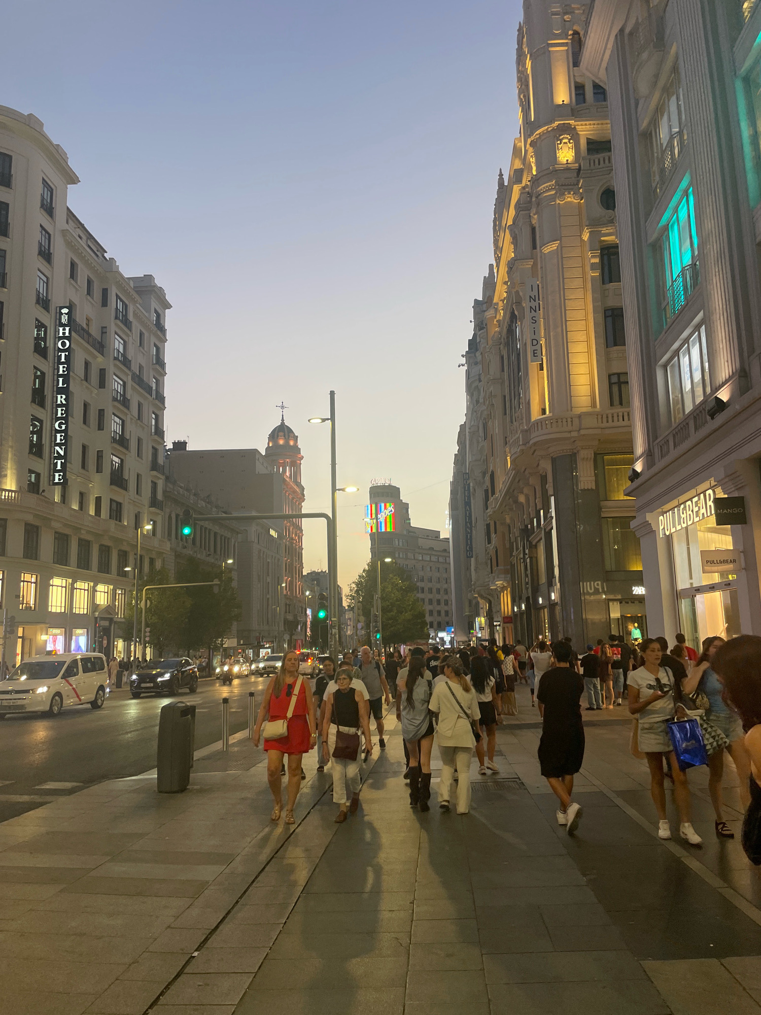 This is a nighttime street scene in Madrid, Spain, on a wide pedestrian boulevard. The sky is a dusky blue, indicating either early evening or late twilight. The street is lined with tall, ornate buildings, illuminated by warm yellow and green lights. One building on the left prominently displays a vertical illuminated sign reading "HOTEL REGENTE." Across the street, a building features a vertical sign for "INNSIDE." Further down the street, shops like "PULL&BEAR" and "MANGO" are visible, their storefronts lit up. A large, multi-colored digital billboard is visible in the distance above a building.
The boulevard is bustling with people walking in both directions on the wide sidewalk. Many individuals are dressed in casual summer clothing. A street with vehicular traffic runs alongside the pedestrian area. Several cars, including a white taxi van and a black SUV, are visible on the street. Traffic lights are also present. Streetlights and lights from the buildings cast shadows on the pavement. The overall atmosphere is lively and suggests a popular urban area.