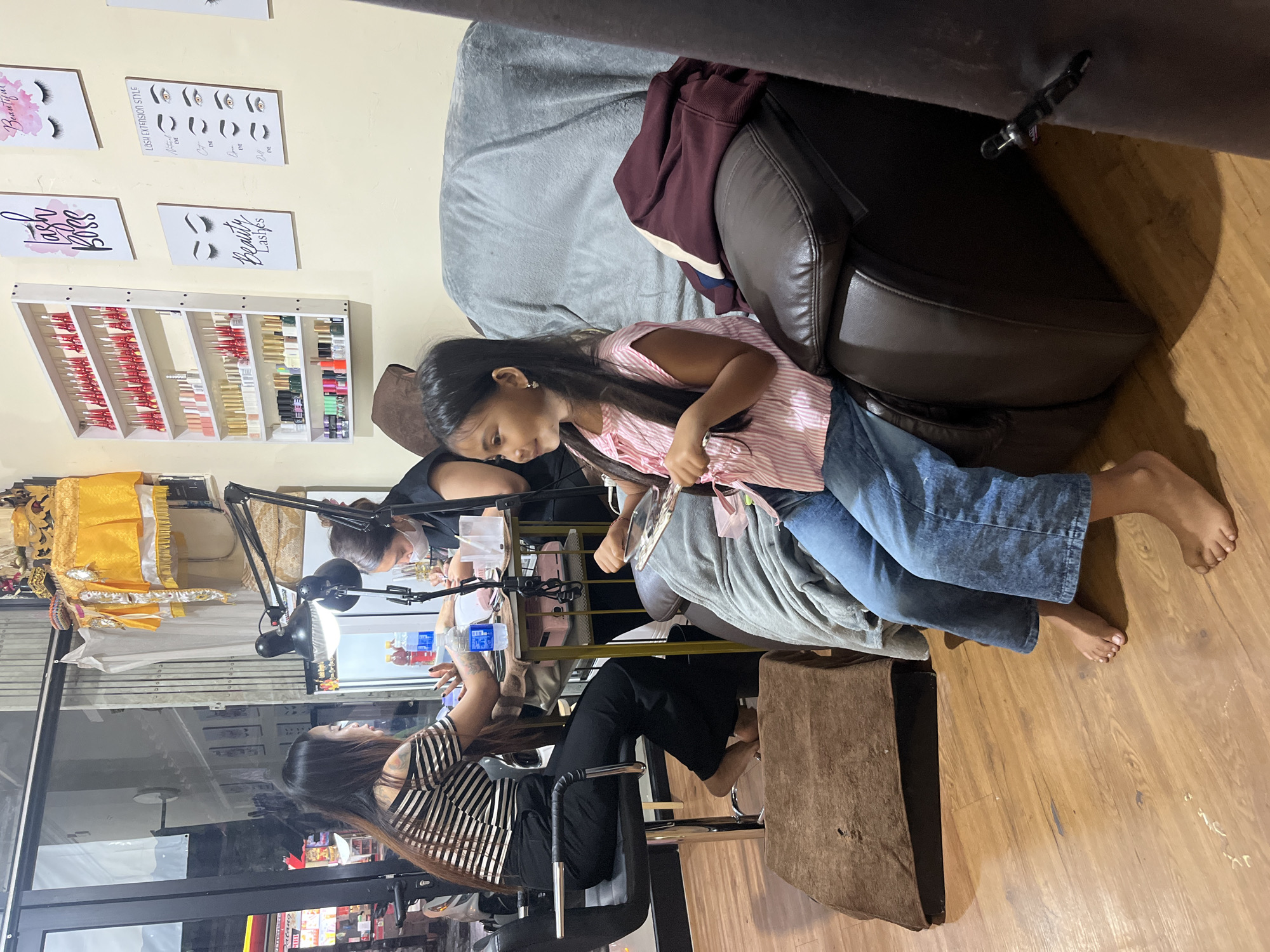 In North Kuta, Indonesia, at a nail salon, two women are receiving or giving manicures. One woman, with long dark hair and a striped off-the-shoulder shirt, is seated on a black chair with her feet resting on a brown footrest, looking down at her hands. The other woman, wearing a black shirt and a white mask, is leaning over a table with a lamp, working on a client's nails. In the foreground, a young girl with long dark hair, wearing a pink and white striped shirt and blue jeans, sits on a brown leather recliner, holding a small mirror and looking to her left. The salon has a shelf displaying numerous bottles of nail polish, and posters of eyelashes are on the wall. The floor is made of light-colored wood.