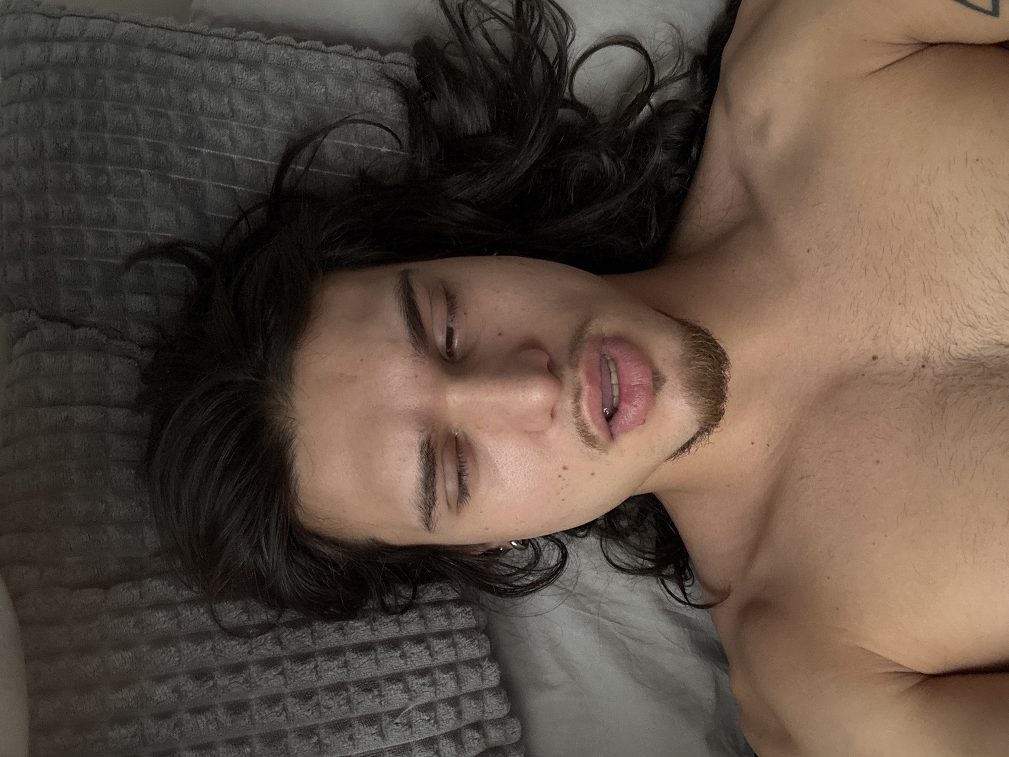 A man lies on a gray textured pillow and a gray sheet. He has long, dark, curly hair. He is shirtless and his face is slightly obscured by the angle. He has a piercing in his lip and an earring in his left ear. The tattoo on the arm is barely visible. The location is unknown.