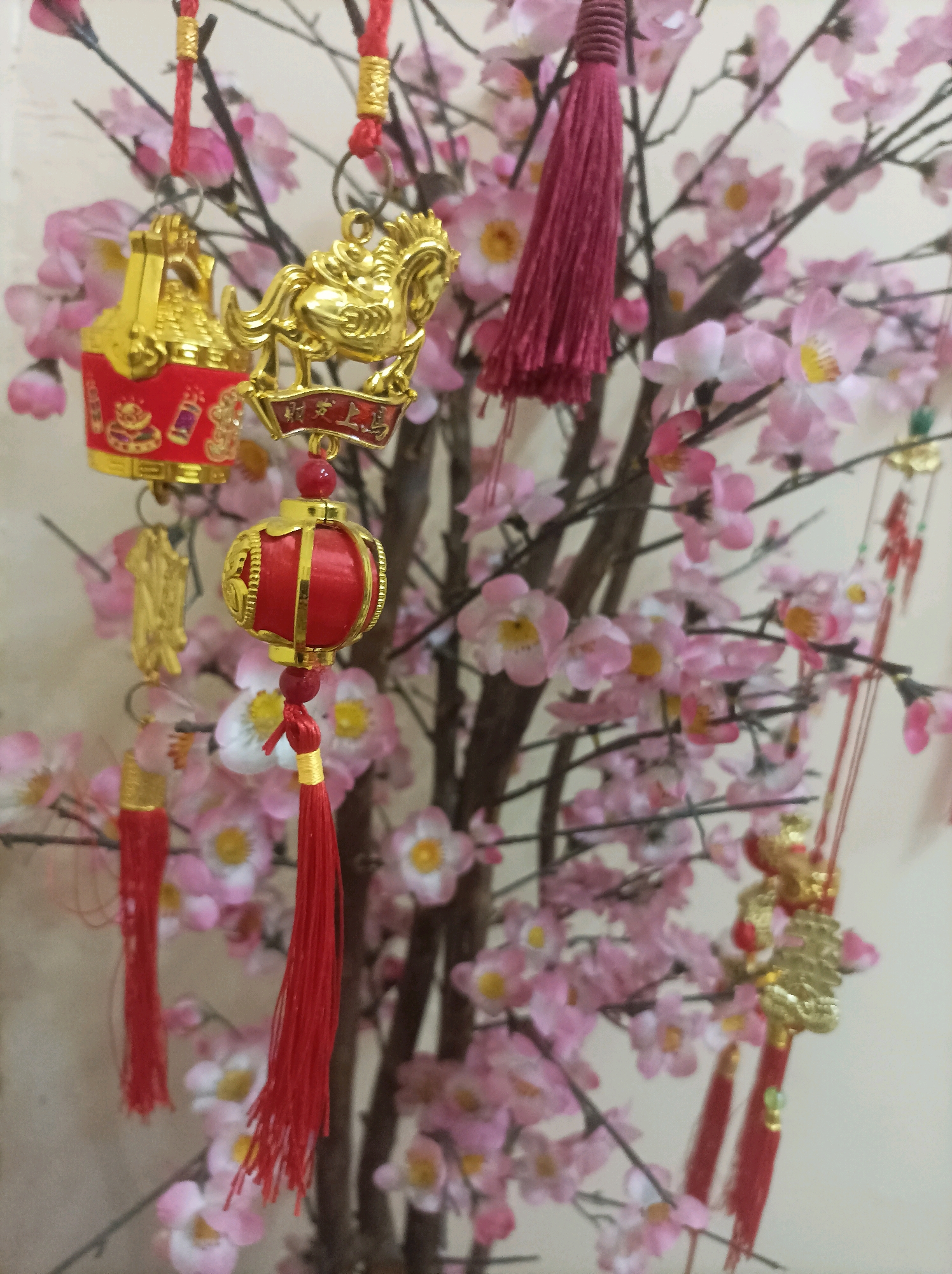 This is a close-up, eye-level shot of a decorative cherry blossom tree with hanging ornaments. The tree is in focus, with its branches laden with pink and white artificial blossoms. Several ornaments are visible, predominantly gold and red. These include a small golden horse, a golden bell, and red lanterns with gold accents. Red tassels are also attached to some of the ornaments and the tree branches. The background is a plain, light-colored wall, suggesting an indoor setting. The lighting is even, indicating it's likely daytime or a well-lit interior. There are no people or animals present. This image does not provide any information to determine a specific location in Bekasi, Indonesia.