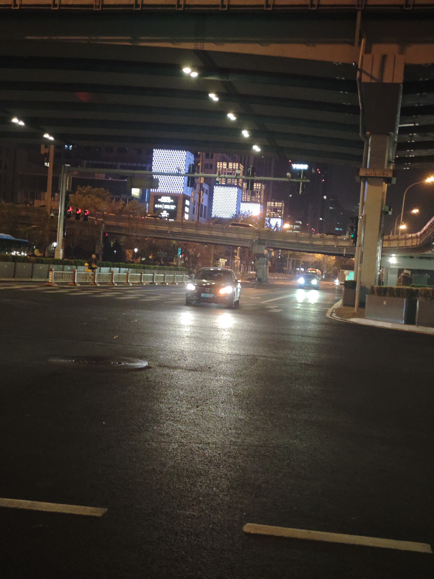 This nighttime photograph captures a dynamic urban scene in Chaoyang District, China, characterized by its multi-layered infrastructure and artificial illumination. The setting is a city intersection at night, with the dark sky overhead and various light sources dominating the view. The road surface appears wet and reflective, suggesting recent rain or damp conditions.
In the foreground, a dark-colored car with bright headlights is approaching the viewer, its lights casting a glow on the wet asphalt. Further down the road, another vehicle is visible, also with its headlights on. On a median or sidewalk area to the left, a person wearing a bright yellow jacket stands behind a low white barrier with orange posts.
The scene is dominated by a massive elevated road or overpass structure that spans across the top of the frame, supported by large concrete pillars. Traffic lights are visible at various points, with some displaying red and others green, indicating active traffic flow. In the background, tall city buildings are illuminated, with one prominent structure featuring a distinctive grid pattern of square lights across its facade, likely an LED screen or elaborate architectural lighting. Faint text in Chinese characters is discernable on some of these illuminated building facades. Faded white road markings are visible on the asphalt, along with a manhole cover in the foreground. The overall impression is one of a bustling, modern metropolis continuing its rhythm after dark.