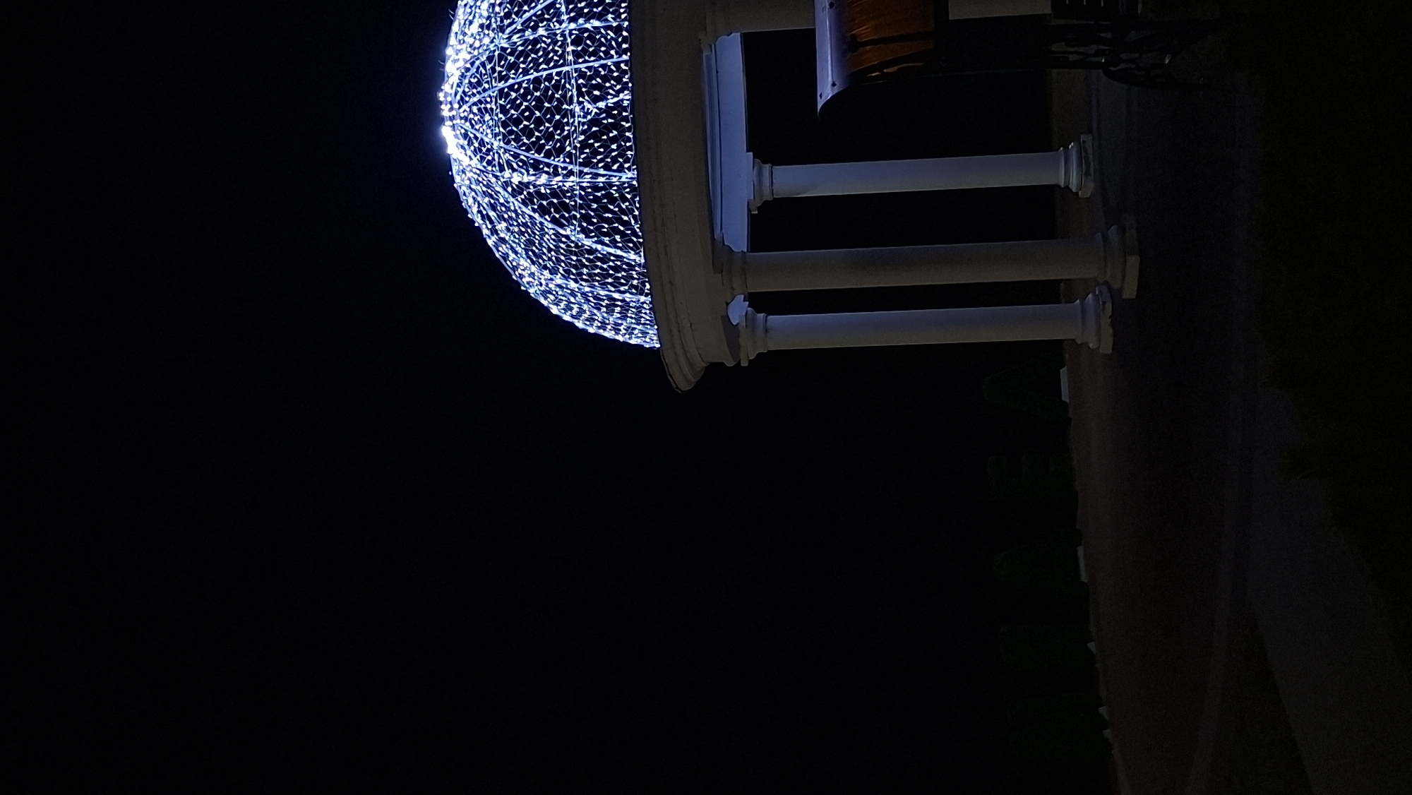 An illuminated dome structure with white lights sits atop a white, classically designed building with columns. The structure is covered in a lattice of white lights, creating a dome shape. To the right of the building, a part of a dark-colored vehicle or cart with an orange top is visible. In the background, large, dark green hedge letters spelling out "MYADZYEL" are visible, illuminated by subtle lighting. The scene is dark, suggesting it is nighttime. The ground in front of the building appears to be paved.