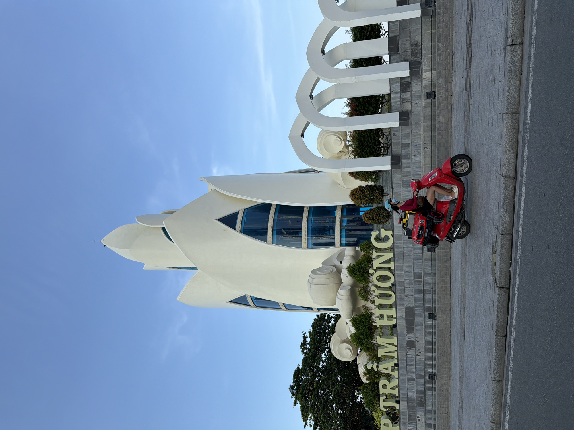 The image displays an urban scene in Nha Trang Ward, Vietnam, centered on the Tháp Trầm Hương (Agarwood Tower). The tower, located in the middle-ground, is a multi-tiered, white architectural structure with an organic, petal-like form, featuring several large blue-tinted glass windows with triangular and curved shapes. At the base of the tower are large, white, scroll-like sculptural elements and green shrubbery. To the right of the tower, a series of five white, segmented arches form an open structure on a raised grey platform. Below the tower, large, light-colored block letters spell "P TRẦM HƯƠNG" against a stepped, light grey stone wall. A large green tree is visible to the left of the tower's base. In the foreground, a person is riding a red motor scooter from left to right on a grey paved sidewalk. The rider wears a blue helmet, a light blue face mask, a dark red long-sleeved top, and black shorts. A red toy jeep is secured upright on the rear rack of the scooter, behind a black bag or container. The street and curb are visible in the immediate foreground. The sky is clear and light blue with a few faint wisps of white cloud. The lighting indicates daytime.
