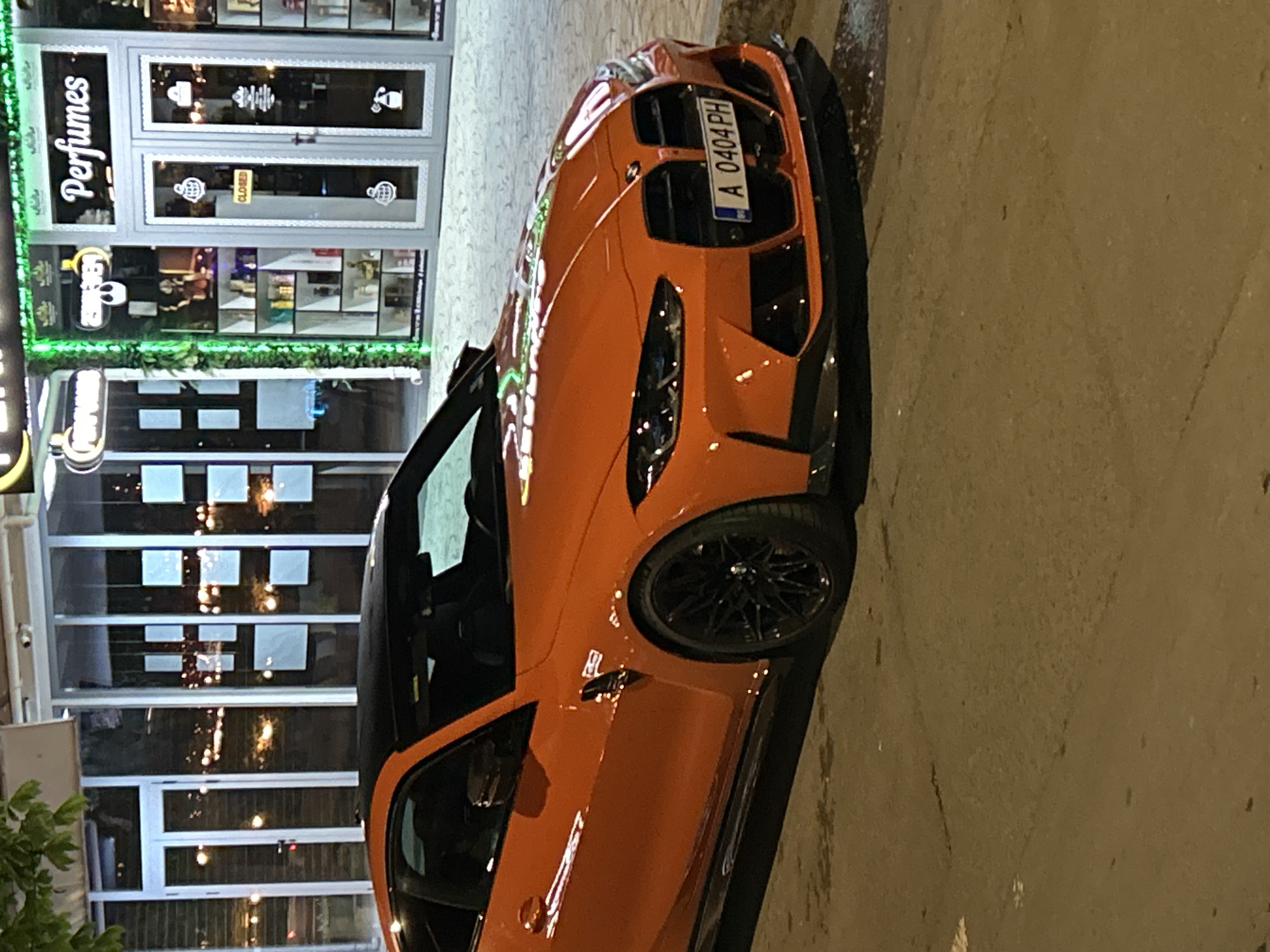 An orange BMW sports car is parked on a street near a closed perfume store. The store has "Perfumes" written in white cursive letters and neon green lights around the store's sign. The car's license plate reads "A 0404 PH". The location is possibly in the city of Nesebar, Bulgaria.