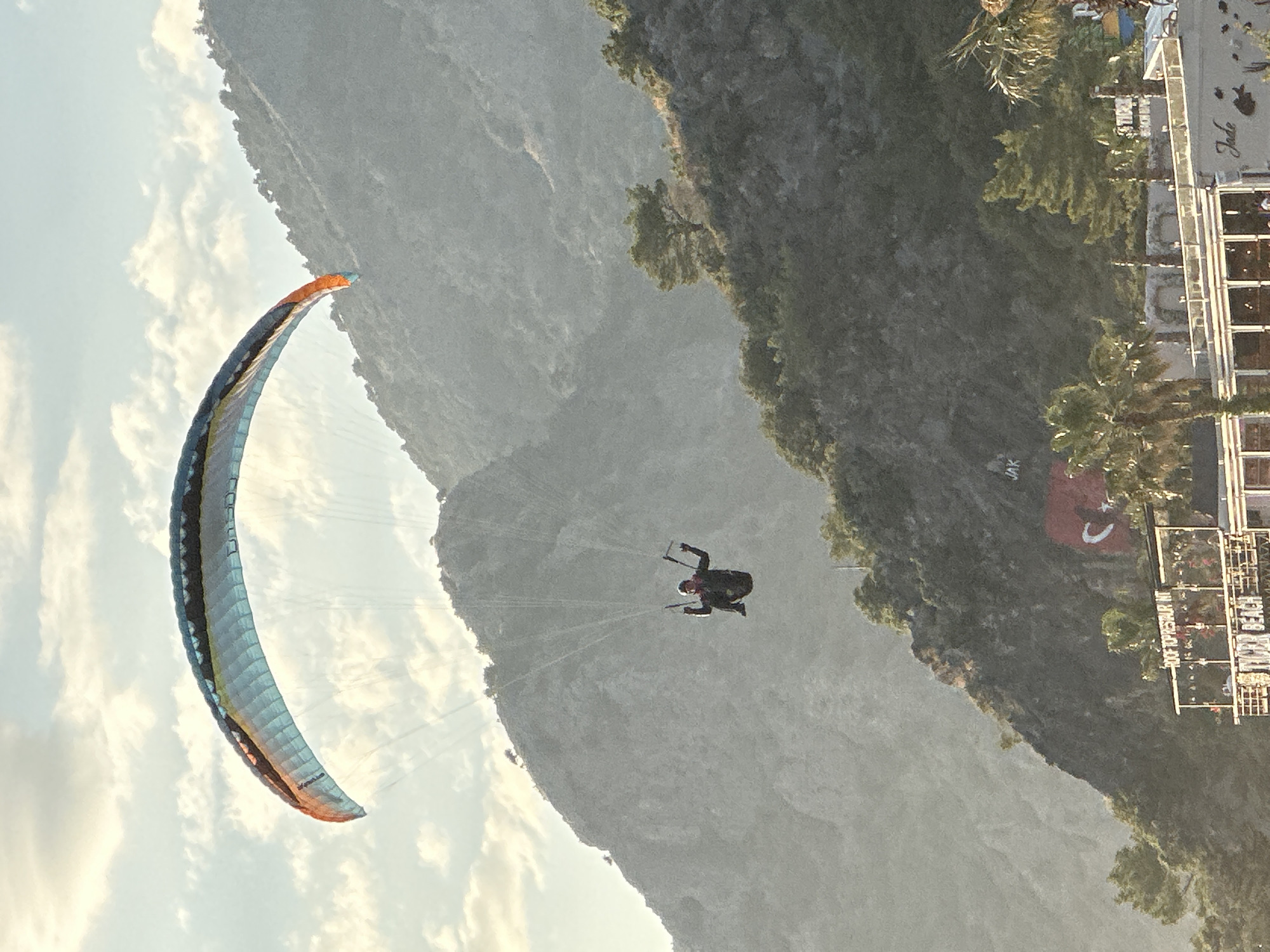 A medium outdoor shot presents a person parasailing against a backdrop of rugged mountains in Fethiye, Turkey. The parasail, predominantly blue with orange accents, is prominently displayed in the left third of the frame. The parasailer, dressed in dark attire, is centered in the middle third of the image. The background features a steep, rocky mountain range. On the right, vegetation-covered hills and an urban area, possibly a town or resort, are visible. A Turkish flag is affixed on the side of the mountain range. The weather appears calm, with some clouds in the blue sky.