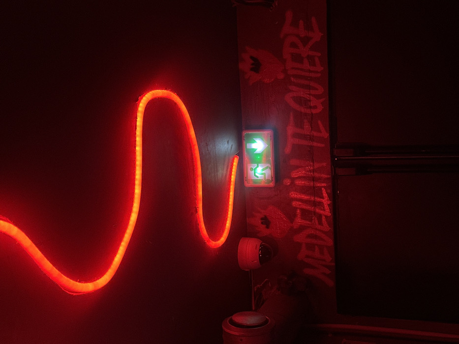 An interior scene, saturated in red light, depicts a ceiling-mounted, sinuous red LED strip. Below, on a dark wall, a white dome security camera is positioned to the left of a rectangular, green-illuminated emergency exit sign, which displays a white running figure and a downward arrow. Below these fixtures, the phrase "MEDELLIN TE QUIERE" is rendered in light-colored, graffiti-style script. Dark, stylized motifs, possibly resembling hearts, are visible near the emergency sign. To the far left, a circular vent or speaker is partially discernible. The lower frame reveals the upper section of dark, vertical panels, likely doors. No individuals are present. The setting is consistent with a commercial establishment, such as a bar or club, located in the city of Paris, France.