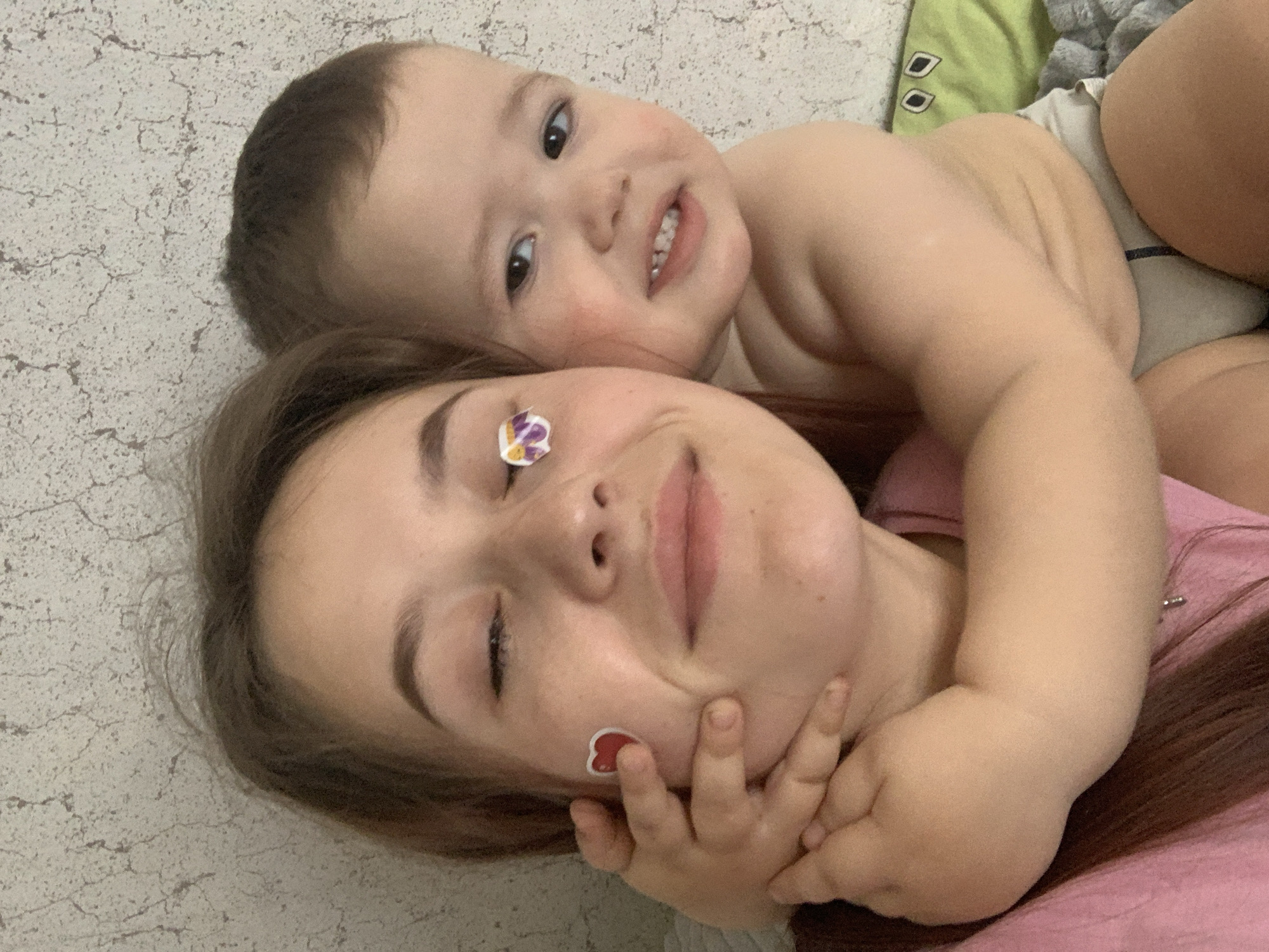 A young child is lying on top of a young woman. The woman is lying on her back, smiling with a flower sticker on her forehead and a heart sticker on her cheek. The child is lying on top of the woman with their arms around her. The background is a textured white surface. The location is unknown and cannot be inferred from the image. The city of Фоки, Russia cannot be inferred from the image.