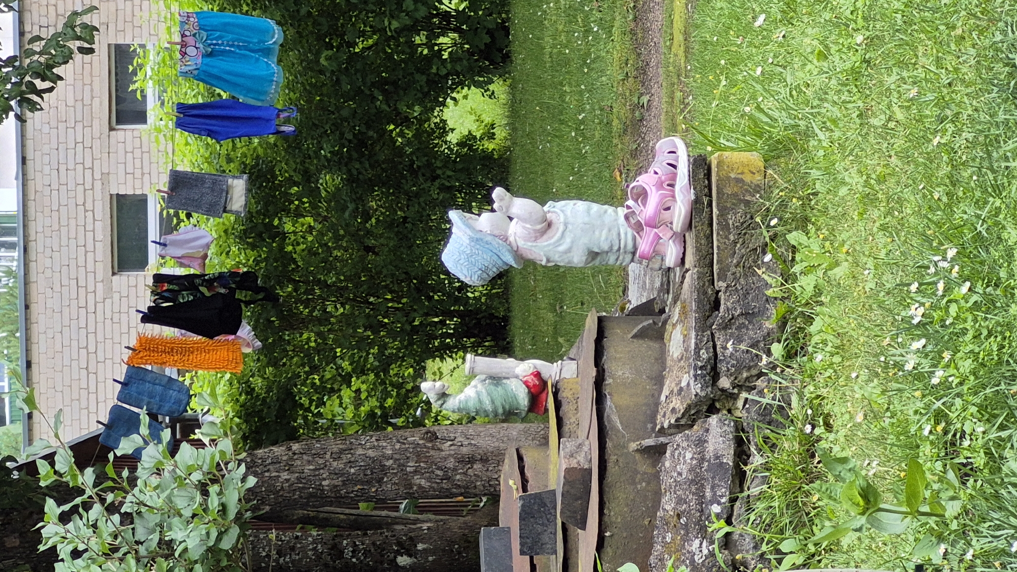 The image depicts a backyard scene in Švenčionys, Lithuania, with laundry hanging on a clothesline in the background, a brick building, and lush green foliage. In the foreground, there are two garden statues. One is a gnome-like figure dressed in green with a red base, standing on a tiered structure. Next to it is a statue of a child wearing a blue hat and light-colored overalls, with pink sandals placed in front of it. The ground is covered with grass and small white flowers.