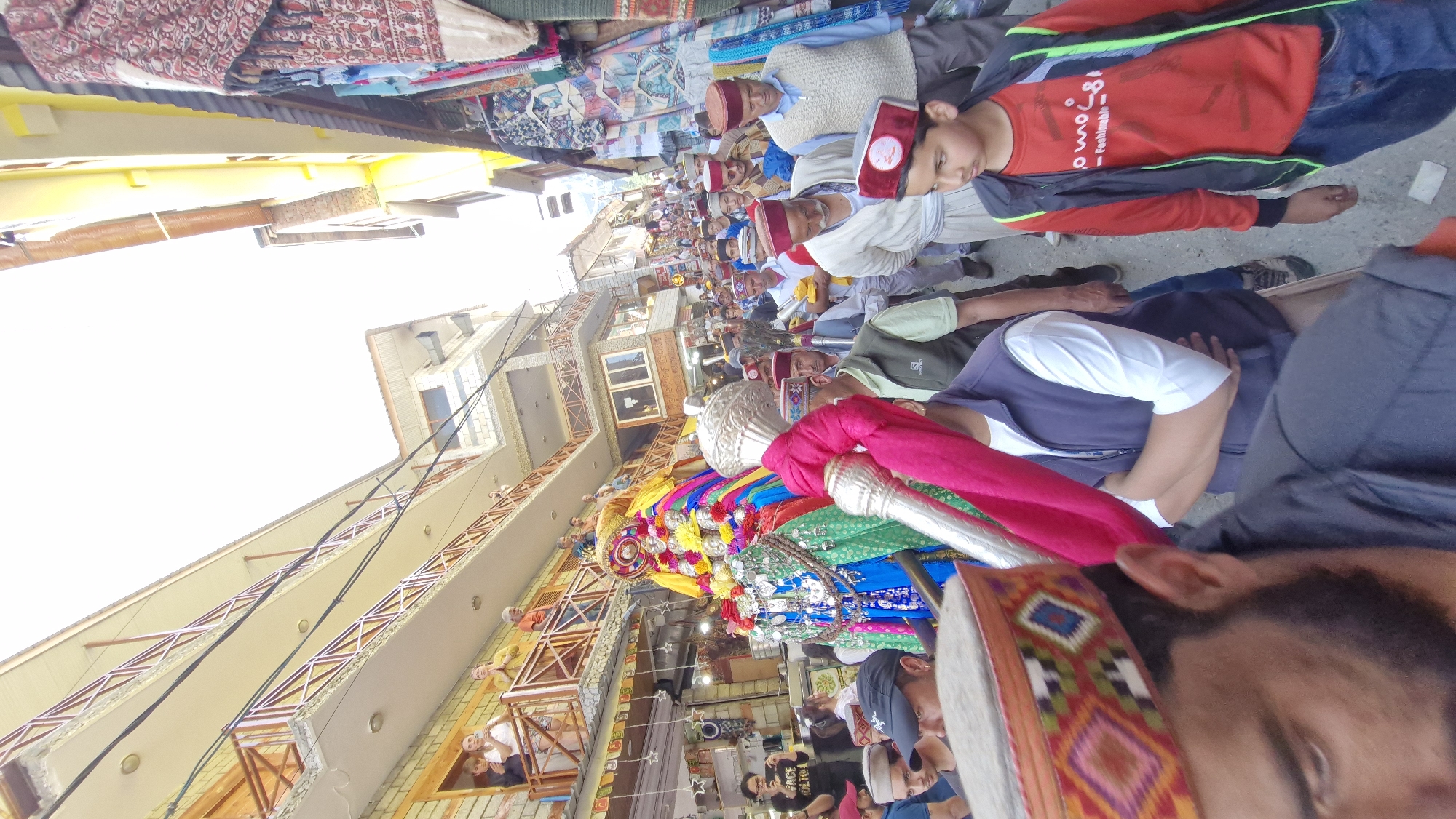 The image captures a vibrant procession in the narrow street of Vashisht, India. A colorful, ornate structure, possibly a deity's palanquin or idol, is the central focus, adorned with bright fabrics, garlands, and decorative elements. It is being carried by several individuals who are partially visible. A large group of men, many wearing traditional Himachali caps with intricate patterns, are walking alongside and behind the palanquin, participating in the event. Some are carrying what appear to be ceremonial items. On either side of the street, buildings with multiple stories line the path, with some balconies occupied by spectators. Shops with displayed textiles and other goods are visible at ground level. The overall atmosphere suggests a lively religious festival or cultural celebration.