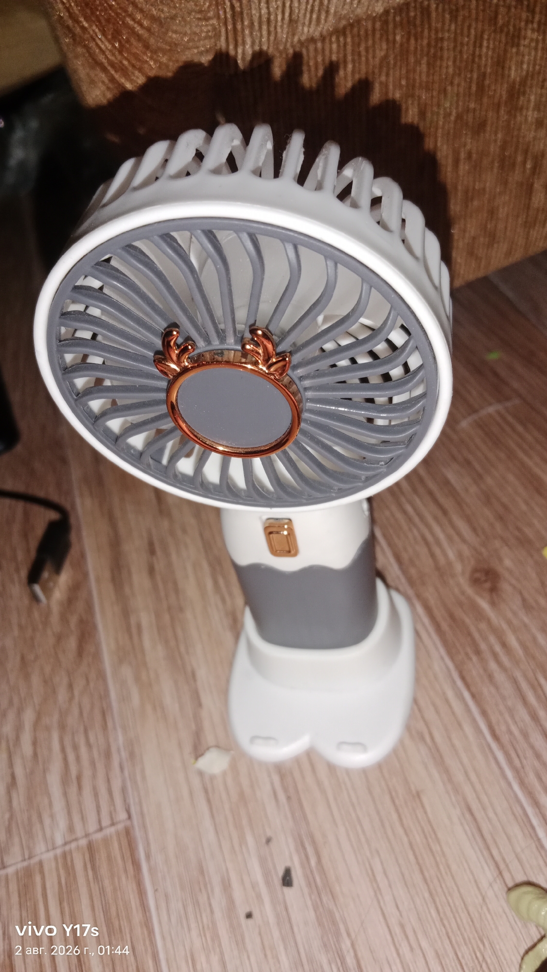 A close-up, eye-level shot shows a white and grey portable fan with a decorative element resembling antlers on the fan guard. The fan is positioned on a wooden floor, with its base facing downwards. A brown, textured surface, possibly upholstery, is visible in the upper portion of the frame, casting a shadow onto the fan. A black USB cable is partially visible to the left of the fan. The scene is indoors, and the overall lighting suggests it is not daytime. The image contains overlaid text at the bottom left corner indicating "vivo Y17s" and a date and time stamp in Russian, which translates to "August 2, 2026, 01:44". This date and time stamp is likely metadata from the camera and not indicative of the actual date or time the photo was taken or the fan's location. Therefore, information regarding the city of Rudny, Kazakhstan, cannot be inferred from the image itself.
