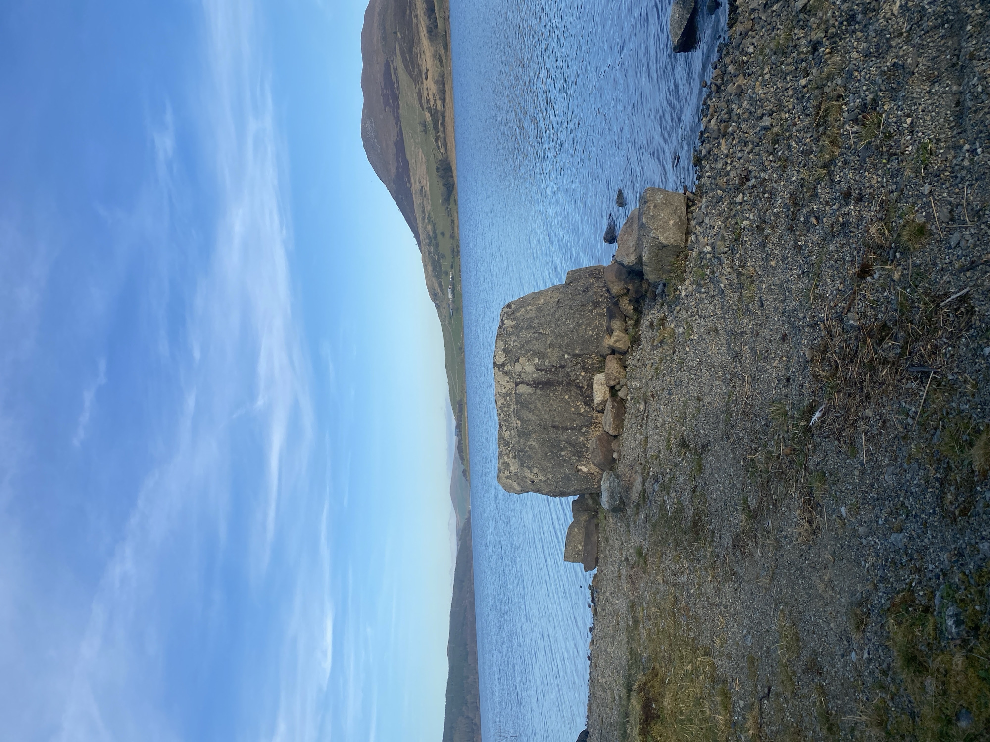 The image depicts a landscape scene in Bala, United Kingdom, showing a body of water, likely a lake, adjacent to a shoreline composed of small gravel and rocks. A large, grey rock sits at the water's edge, with smaller stones nearby. The water surface has a slight ripple effect. In the background, a mountain range is visible on the opposite side of the lake, with varying shades of brown and green indicating vegetation. The sky above is blue with wispy, scattered clouds.