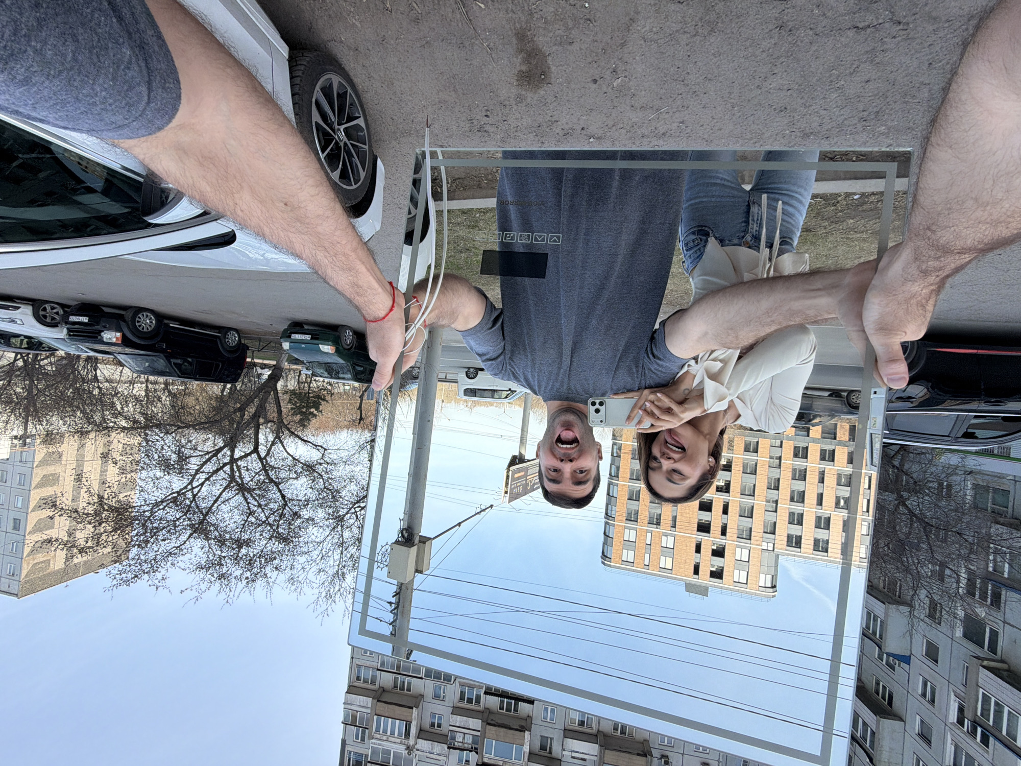 A man and woman are taking a selfie, reflected in a large rectangular glass pane being held up by two arms. The man is wearing a grey t-shirt and is open-mouthed. The woman is in a white blouse and has her tongue out. The background reflection shows two buildings and a clear sky. Vehicles and a leafless tree are also visible. The arms holding the glass pane are fair-skinned and hairy, one with a red string bracelet. The location is in the city of Krasnoyarsk, Russia.