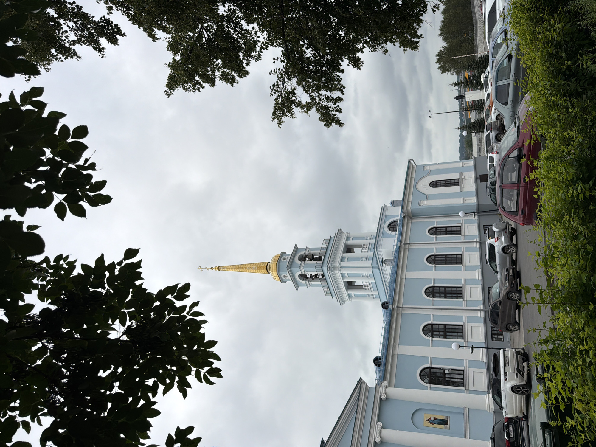 The image shows a light blue building with white architectural details and a tall, golden-domed bell tower. The building appears to be a church or a similar religious institution. In front of the building is a parking lot with several cars. Trees are visible on the left and right sides of the image, with their leaves creating a frame for the scene. The sky is overcast. The location is the Votkinsk City Museum and Exhibition Center in Votkinsk, Russia, which is located on the former territory of the Votkinsk Ironworks. This building complex is a historical landmark in the city.
