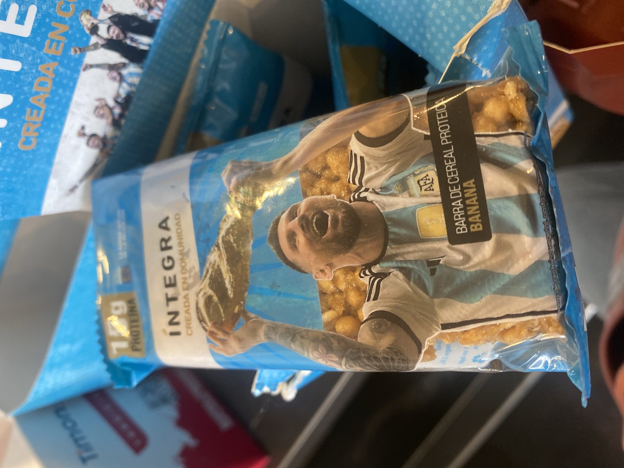The image shows a close-up of a packaged cereal bar featuring a picture of Lionel Messi. The bar has "INTEGRA" printed on it, along with "15g PROTEINA" and "BARRA DE CEREAL PROTEICA BANANA". Messi is depicted wearing an Argentinian football jersey and holding a trophy, with celebratory figures in the background. The packaging is predominantly blue and white, with some red and yellow elements. The background is blurred, but appears to be indoors, with other packaged items visible. There is no discernible information about the specific location within the city of Cordoba, Argentina.