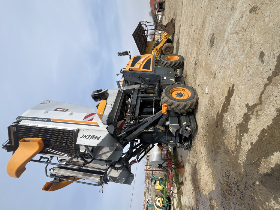 The scene depicts an outdoor agricultural machinery storage or maintenance yard in the city of Terziysko, Bulgaria, under a partially cloudy blue sky.
The primary subject is a large, self-propelled Pellenc grape harvester, predominantly grey, orange, and black, positioned centrally. The machine's harvesting head is elevated, exposing vertical internal rollers, and displays "PELEENC" and "SELECTIV PROCESS" branding. It features large, multi-lugged black tires with orange hubs.
To the right, a yellow JCB backhoe loader or wheel loader, equipped with a front bucket, is parked beneath an open-sided shed with a corrugated metal roof. A red tractor is partially visible further to the right.
To the left of the grape harvester, various agricultural implements are visible, including green and yellow spraying equipment and a red plow attachment. Behind these, a large, horizontal, silver cylindrical tank is present.
The ground is an unpaved surface of dirt and gravel, marked with several dark, irregular stains consistent with pooled water or oil. No individuals are visible within the frame; all machinery appears stationary.