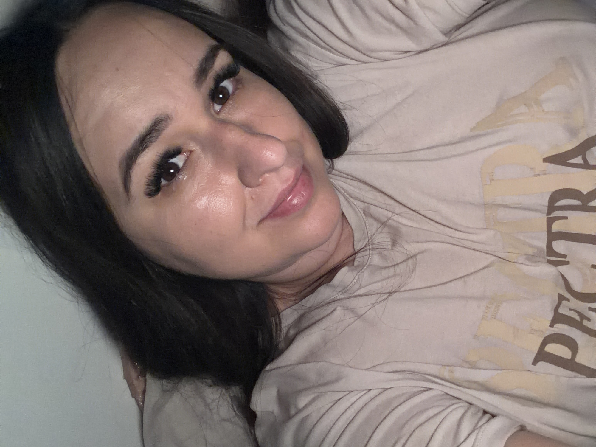 A selfie of a woman lying down, with her head slightly tilted and her body angled towards the viewer. She has dark hair and is wearing a light-colored t-shirt with text on it. The background is a plain wall.