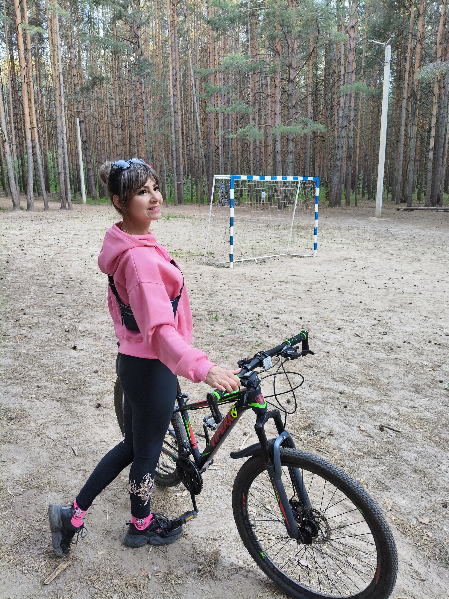 The image shows a woman standing next to a bicycle in a wooded area that appears to be a park or recreational space. The woman is wearing a pink hoodie, black leggings with a design, and black sneakers with pink socks. She has sunglasses on her head and her hair is styled in a bun. She is holding the handlebars of the bicycle, which is black with green accents. In the background, there are tall trees, possibly pine or fir, and a soccer goal with blue and white stripes. The ground is sandy and covered with pine needles. The location is near the city of Chaikovsky, Russia.
