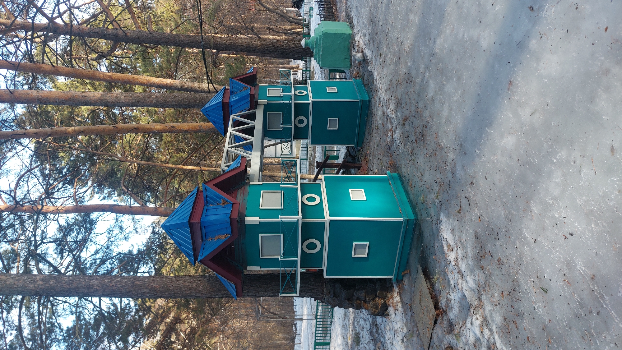 The image depicts a colorful, multi-story structure resembling a whimsical building in a park or recreational area. The structure is predominantly teal with white trim and features windows and circular decorative elements. It has a blue roof with red accents and is connected by white structural supports.
The structure is situated in an outdoor setting with tall pine trees in the background. There's evidence of winter or cold weather, with patches of ice and snow on the ground. The scene appears to be a park or public space, potentially a children's playground, given the nature of the structure.
The location is Novosibirsk, Russia.
