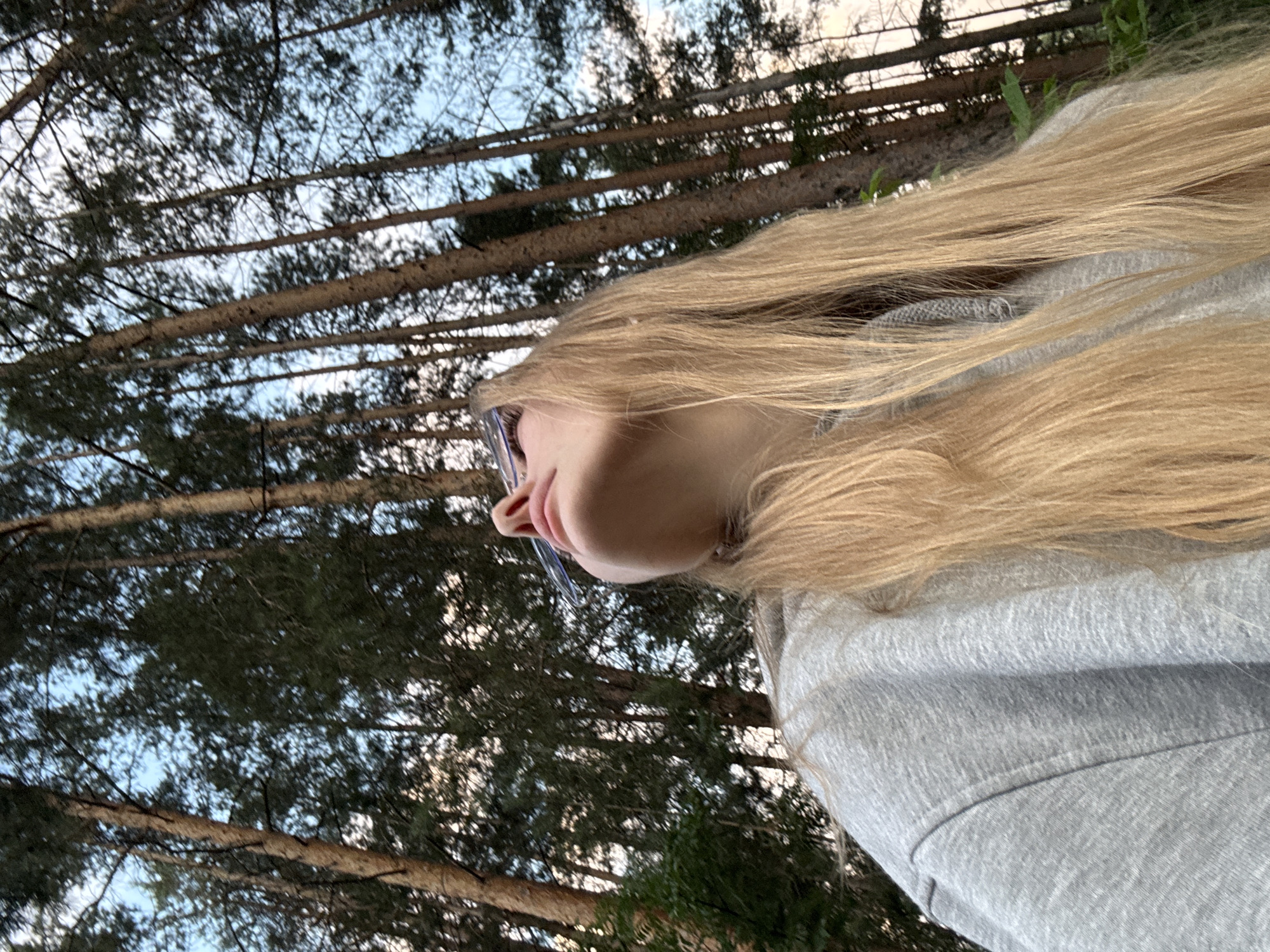 A low-angle, medium shot captures a blonde woman in a grey hoodie looking upwards in a forest. She is wearing clear-framed glasses. The background consists of tall, slender pine trees and a hazy sky. The overall scene suggests an outdoor setting, possibly a park or woodland area within or near the city of Perm, Russia, given the commonality of pine forests in the region. The lighting indicates it is likely late afternoon or early evening.