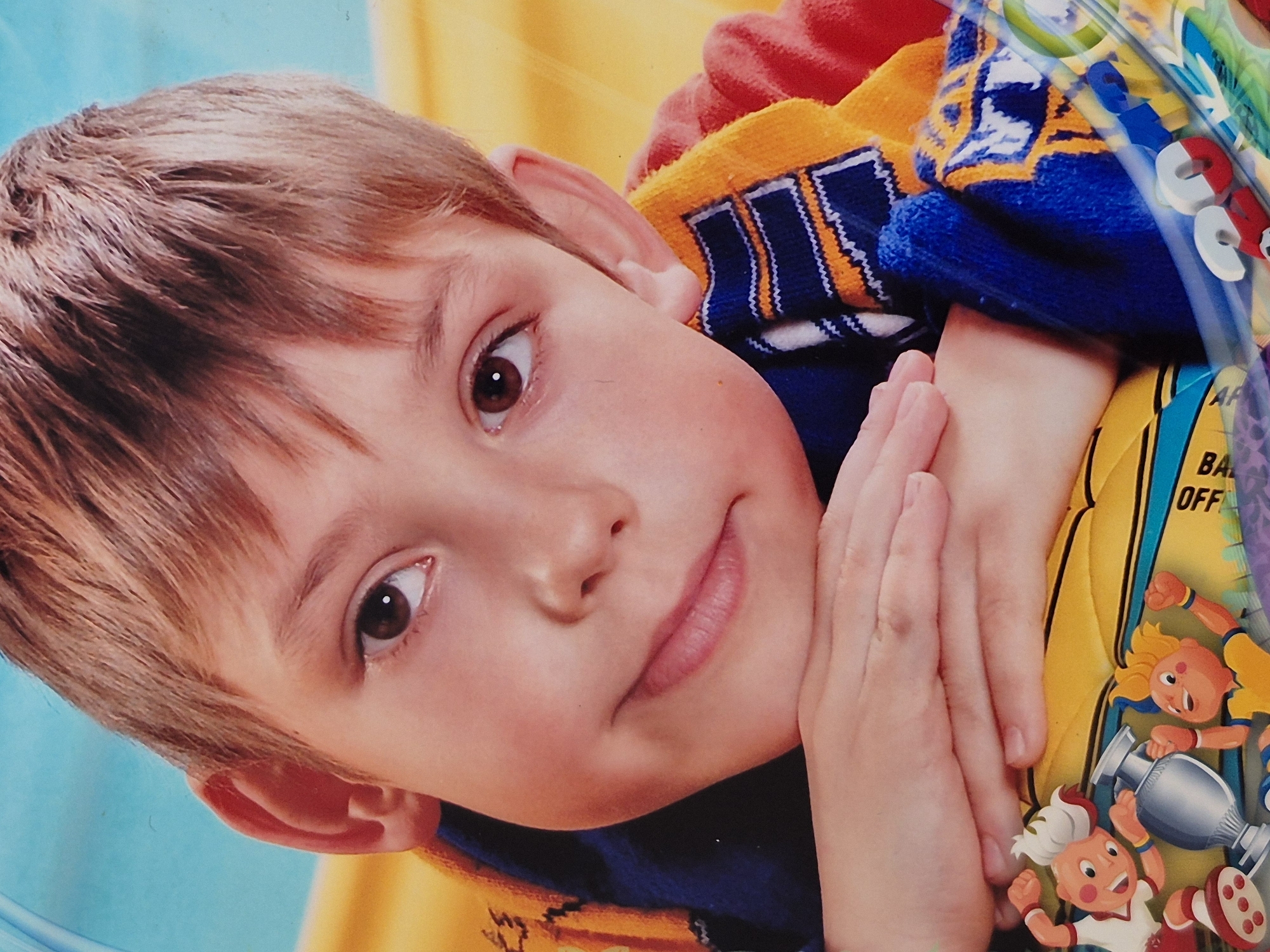 This is a close-up, head and shoulders studio portrait of a young boy with light brown hair and brown eyes, resting his chin on his folded hands. He is wearing a blue and orange striped scarf around his neck, with hints of a red shirt visible. The background consists of a bright blue sky and a yellow surface, suggesting an indoor setting or a backdrop. Part of a yellow ball with cartoon characters and a trophy on it is visible at the bottom of the frame. The image appears to be taken in Schwäbisch Gmünd, Germany, possibly at a photo studio.