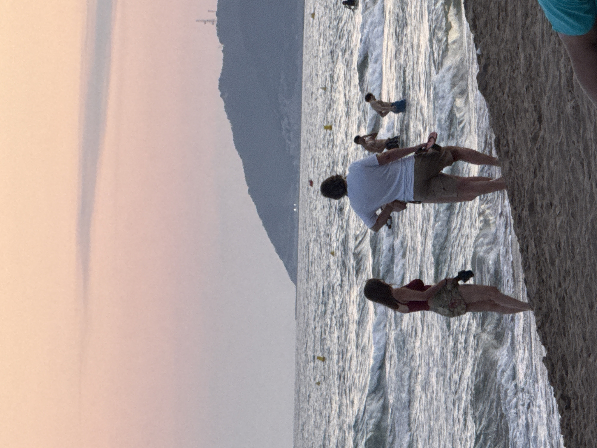 This image depicts a beach scene in Castelldefels, Spain, likely during the late afternoon or early evening, given the soft, warm light. The image is oriented vertically, showing a view of the sea from the sandy shore.
In the foreground, a woman in a red swimsuit and patterned shorts is seated on the sand, looking down at her phone. Behind her and slightly to the left, a man in a white t-shirt and khaki shorts is also seated, holding a phone in his hand and looking to his right. Further out in the shallow water, several other people are visible. One man is standing in the water, while others are further away, some appearing to be swimming or wading. The sea is a light, shimmering blue with visible waves. Small yellow buoys are dotted in the water.
In the background, a large, dark, mountainous landscape rises up, partially obscured by a hazy sky that transitions from a soft pink to a pale grey. The texture of the sand is rough and granular. The overall atmosphere appears relaxed and casual, with people enjoying a day at the beach. There is no visible text in the image.