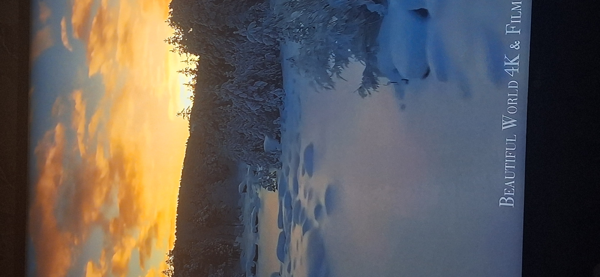 The image shows a tranquil winter landscape at sunrise. The sky is ablaze with hues of orange and yellow, indicative of a rising sun, with scattered clouds adding texture to the vibrant sky. Below the sky, a dense forest of pine trees, their branches laden with snow, forms a dark silhouette against the bright horizon. In the foreground, a snow-covered plain stretches out, dotted with irregular shapes of snow-covered rocks or mounds. A frozen body of water, possibly a lake or river, is visible to the left, also covered in a thick blanket of snow. The overall impression is one of serene, untouched natural beauty. The text "BEAUTIFUL WORLD 4K & FILM" appears in the bottom right corner of the image, suggesting it is a still from a nature documentary or a visually stunning film. There is no indication of the city of Bila Tserkva, Ukraine, in the image.