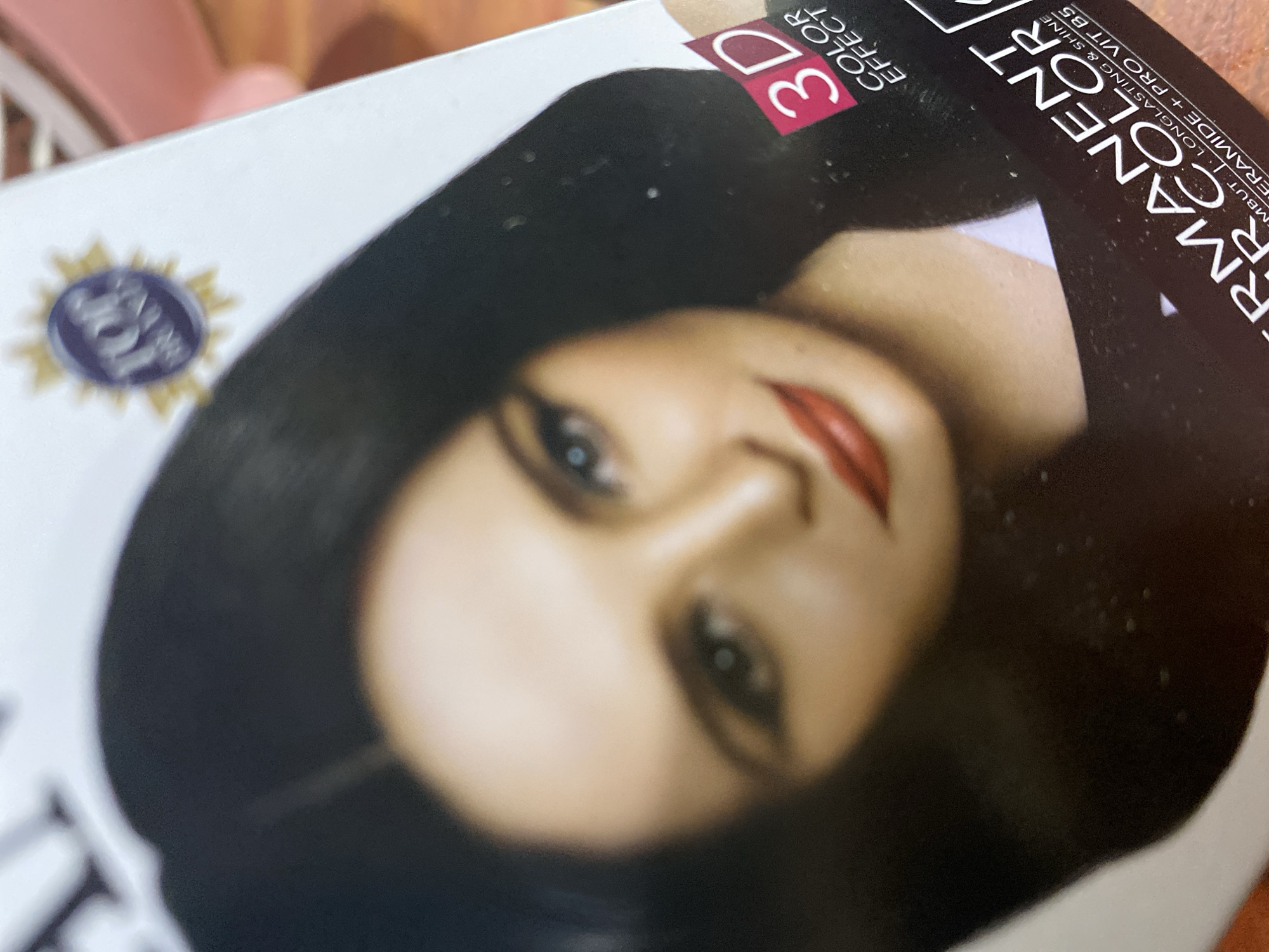 The image is a close-up of a hair dye box featuring a woman's face and black hair. The box has text, including "3D COLOR EFFECT" and "PERMANENT COLOR," along with some smaller text that is partially legible. A circular logo with "TOP" in it is visible on the upper right portion of the box. The image is taken from a slightly elevated angle, suggesting it is a product displayed on a surface, possibly in a retail environment within Ta', Indonesia, although the specific location within the city is not identifiable from the image.