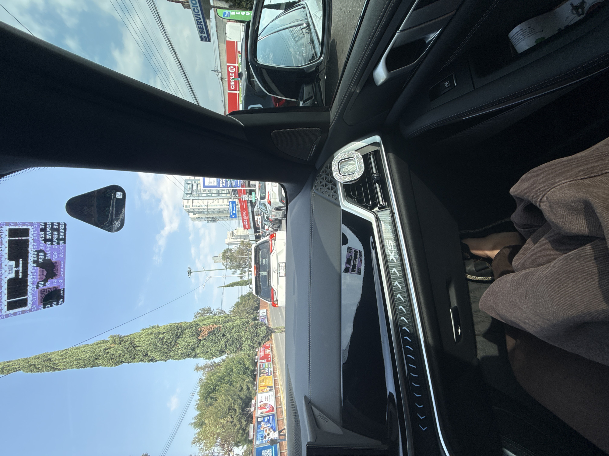 Here is a detailed description of the image:
**Environment:**
The image appears to be taken from inside a car, likely the driver's perspective. Outside, it's a bright day in what seems to be a bustling urban area. There are buildings visible in the background. Tall trees are present along the road.
**Objects & Subjects:**
* **Car Interior:** The focus is on the car's interior, showing the dashboard, air vents (one with a bejeweled decorative object), the side mirror, and the driver's side window. The car model appears to be a BMW X5.
* **Car on the Road:** There is a white vehicle with license plate TTB-395-A ahead.
* **Roadside:** The image shows a Circle K store and multiple billboards.
* **Person:** Part of a person wearing a brown jacket is visible in the passenger seat.
**Location (Based on Visual Clues):**
The presence of a Circle K store along with the license plate information suggests that this image was taken in Puebla, Mexico. The visual details of the buildings and street layout also align with Puebla.