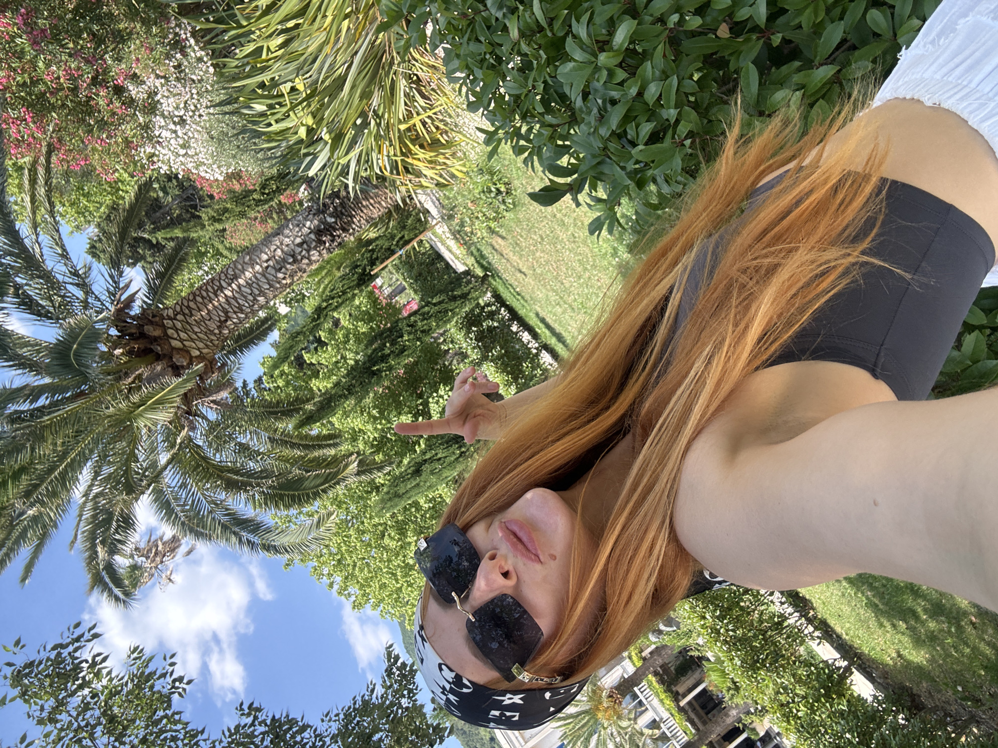 A young woman with long, auburn hair is taking a selfie outdoors. She is wearing large, black sunglasses and a black and white bandana tied around her head. She is wearing a dark grey sports bra and a white skirt. Her right hand is raised, with her index finger pointing upwards. She is standing in a garden with lush green foliage, palm trees, and blooming flowers. In the background, a white building with a white railing is visible. The sky is blue with white clouds. The scene appears to be in a warm, sunny climate. The location is the city of Budva, Montenegro.