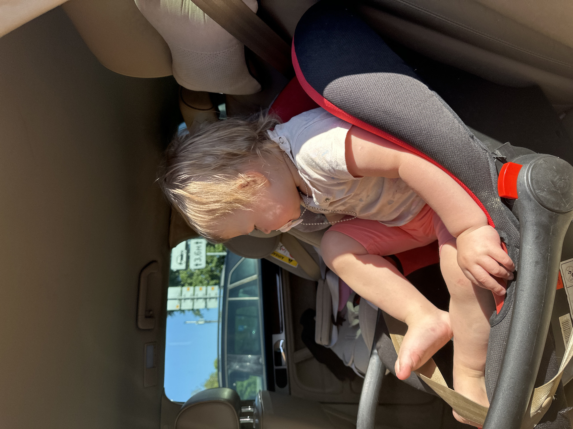 A young child, approximately 1-3 years old, with short blonde hair, is seated in a child safety car seat in the rear of a vehicle. The child wears a light-colored short-sleeved polo shirt and coral-colored shorts, and is barefoot. A white pacifier is in the child's mouth, connected by a silver-colored beaded chain necklace. The child is turned slightly to the right, facing away from the camera. Strong sunlight illuminates the child's left side.
The child safety seat is primarily dark gray with a red border on the left side and a red buckle release button at the crotch strap. Beige harness straps are visible, with one over the child's right shoulder and another between the child's legs, indicating the child is secured. The interior of the host vehicle features light-colored (beige/tan) headrests and ceiling. A dark-colored door panel with a window and handle is visible on the left.
Through the vehicle's window, an urban road environment is visible. A clear blue sky is overhead. A dark-colored SUV or wagon is partially visible in traffic further down the road. A prominent white rectangular road sign with a black border displays a black car icon and the text "↑ 3,6m", indicating a maximum height restriction of 3.6 meters ahead. To the left of this sign, a pole supports several vertically stacked white rectangular signs, which are indistinct due to distance. Green trees and bushes line the roadside. While the signage format using "3,6m" (comma as decimal separator) is consistent with Russian road signage, no specific landmarks, street names, or unique architectural features are discernible to precisely identify the exact location within Nizhny Novgorod, Russia. The scene depicts a typical urban roadside environment.
