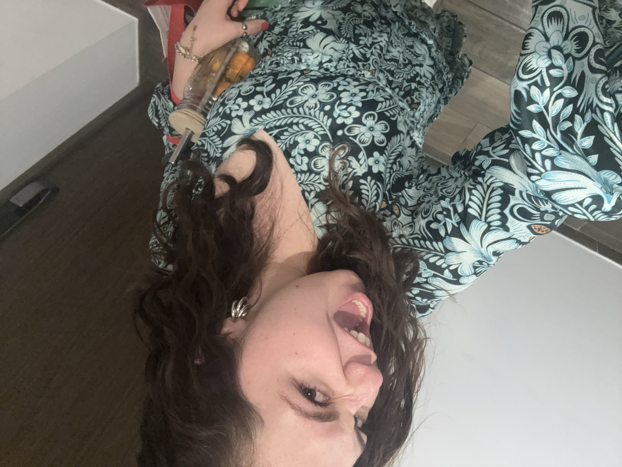 A woman with curly brown hair, fair skin, and an open mouth is pictured in this portrait-style image. She is wearing a patterned dress with a teal and dark floral design. She is holding a glass jar containing orange objects. The location is potentially indoors, based on the neutral-colored walls. The information available does not allow to determine if the scene is in Mexico City.