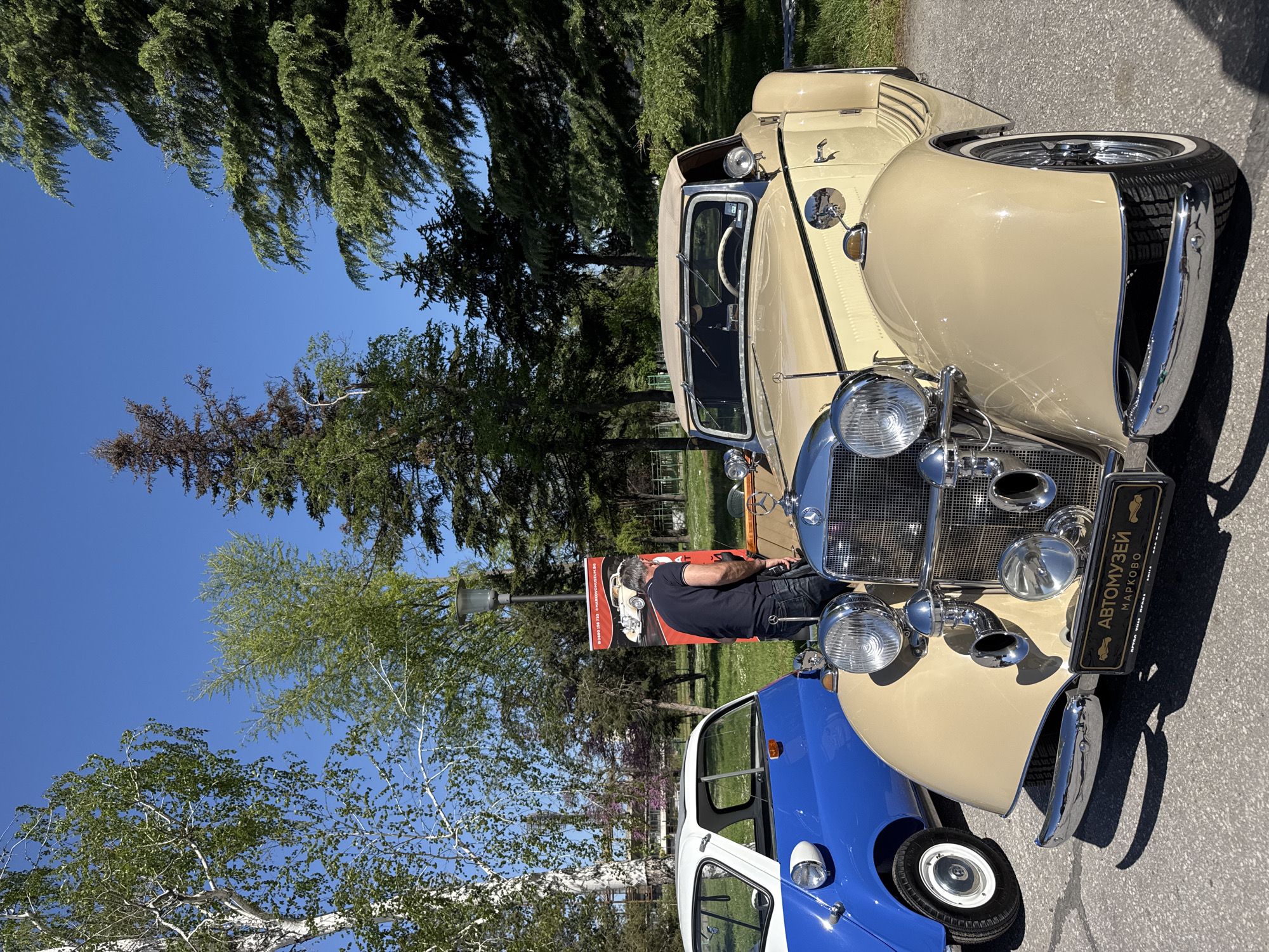 The image shows two classic cars on display in an outdoor setting, likely in Plovdiv, Bulgaria, given the Cyrillic lettering on the license plate identifying an automobile museum in Markovo (near Plovdiv). The car in the foreground is beige and has a chrome grille with vintage-style headlamps. The other car is blue and white. A man in a dark shirt and jeans stands between the two cars, examining a sign. The background includes trees and a clear blue sky. The cars are parked on an asphalt surface. The image is taken during daylight.