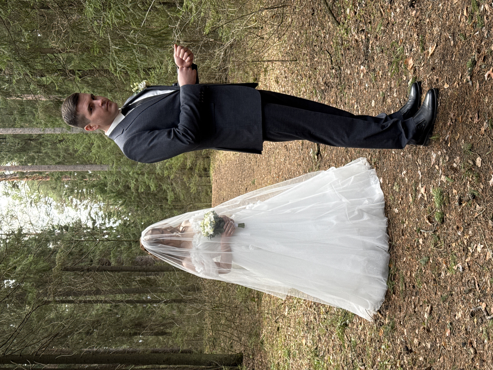 A wedding photograph shows a groom and bride in a forest setting, perhaps near Chaikovsky, Russia. The groom wears a dark suit with a white boutonniere, a white shirt, and a black tie. He has short brown hair and is looking to the left side of the frame while holding his hands near his chest. He is wearing black shoes. The bride wears a white wedding gown covered by a sheer veil. She is holding a bouquet of white flowers and greenery in her hands. The forest floor is covered with brown fallen leaves and bits of grass and other foliage. Tall evergreen trees are visible in the background.