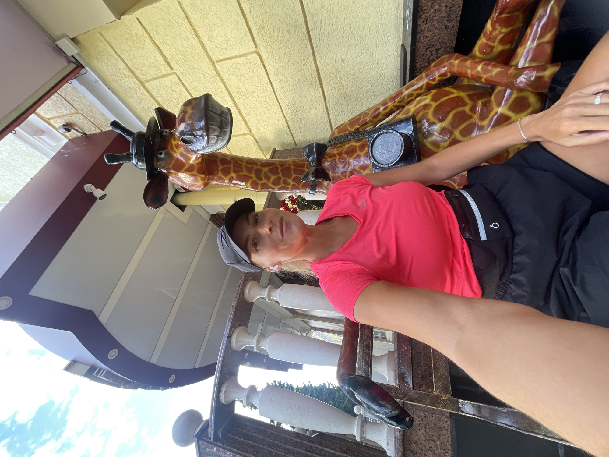 A woman wearing a pink t-shirt, black shorts, a black cap, and wireless earbuds is sitting on a bench next to a large, colorful giraffe statue. The giraffe is wearing a black top hat and a black bowtie. The woman is holding a phone in her right hand, taking a selfie. The background shows a building with yellow textured walls and a white railing. The image appears to be taken outdoors, possibly in a park or public area.