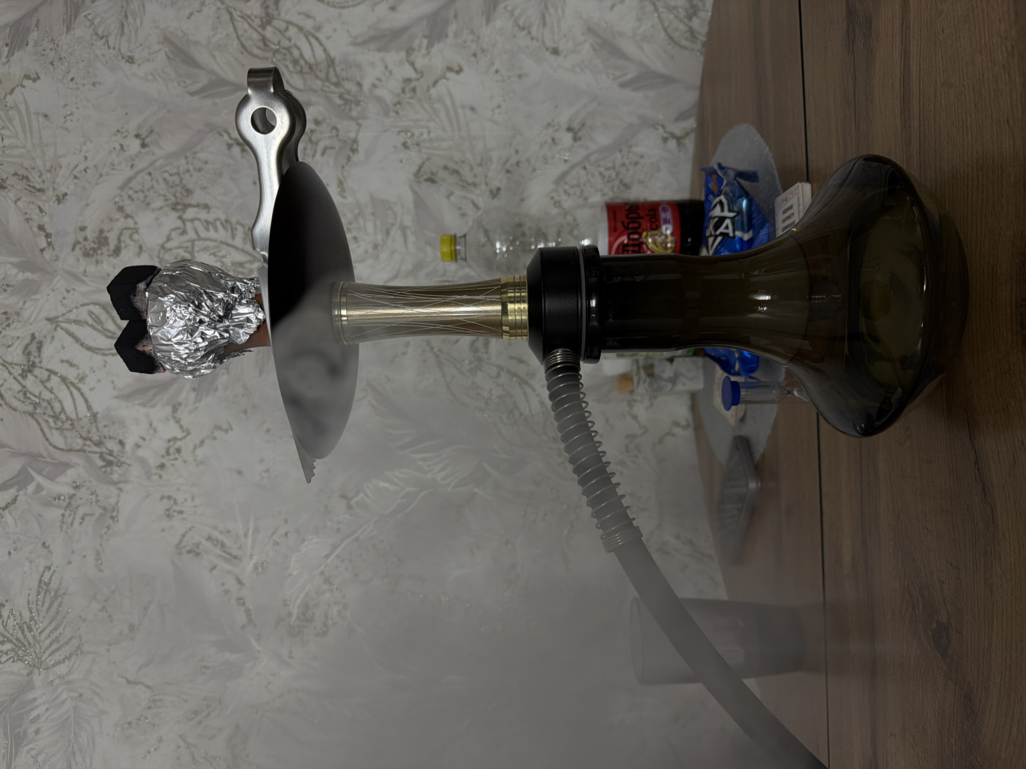 A lit hookah is set up on a wooden surface in a room. The hookah is emitting smoke. The hookah has a glass base and a hose attached. The bowl is covered in aluminum foil and has hot coals on top, held by tongs. In the background, there are various items, including bottles and snack packaging. The surface the hookah is resting on transitions from a patterned white surface to a wooden surface. The location is not evident from this image alone, and therefore, its location in Reshetnikovo, Russia, cannot be determined from this photograph.