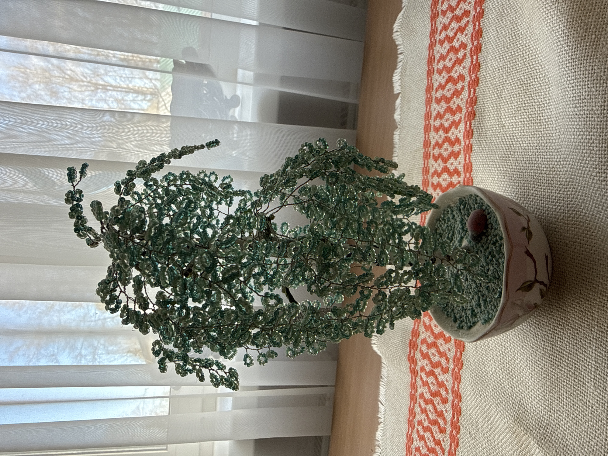 A medium shot depicts a bead tree in a decorative pot on top of a linen cloth. The tree is composed of numerous branches made from wire strung with small, translucent green beads, suggesting foliage. The pot is light-colored, with a hint of floral decoration, and filled with green filler material, likely small pebbles or beads. A single round, dark red or brown object sits on top of the filler. The linen cloth beneath the pot has a beige base with a band of intricate, red-and-white geometric patterns running along its edge. In the background, there are white window blinds, slightly open, admitting natural light. The environment is indoors, likely in a home setting. No people are visible in the frame. There is no evidence suggesting this image was taken in Адам, Russia.