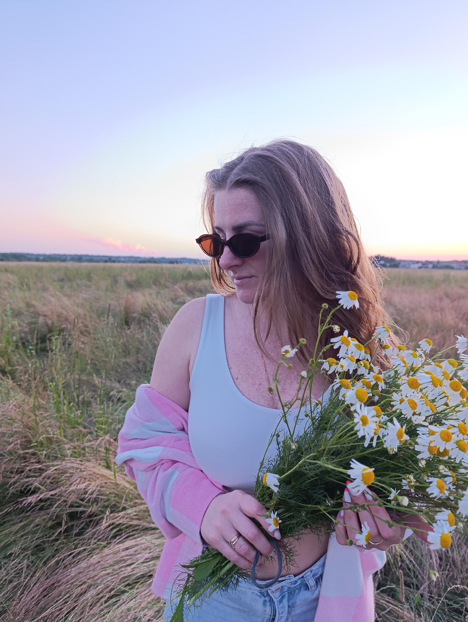 A woman stands in a field of tall, dry grass at dusk. She is holding a bouquet of daisies, their white petals and yellow centers contrasting with the muted tones of the landscape. The woman is wearing a white tank top, a pink and white striped cardigan draped over her shoulders, and blue denim jeans. She has on dark sunglasses and her long, brown hair is loose. The sky above is a soft gradient of pink and blue, suggesting the sun has recently set. The overall mood is peaceful and serene.