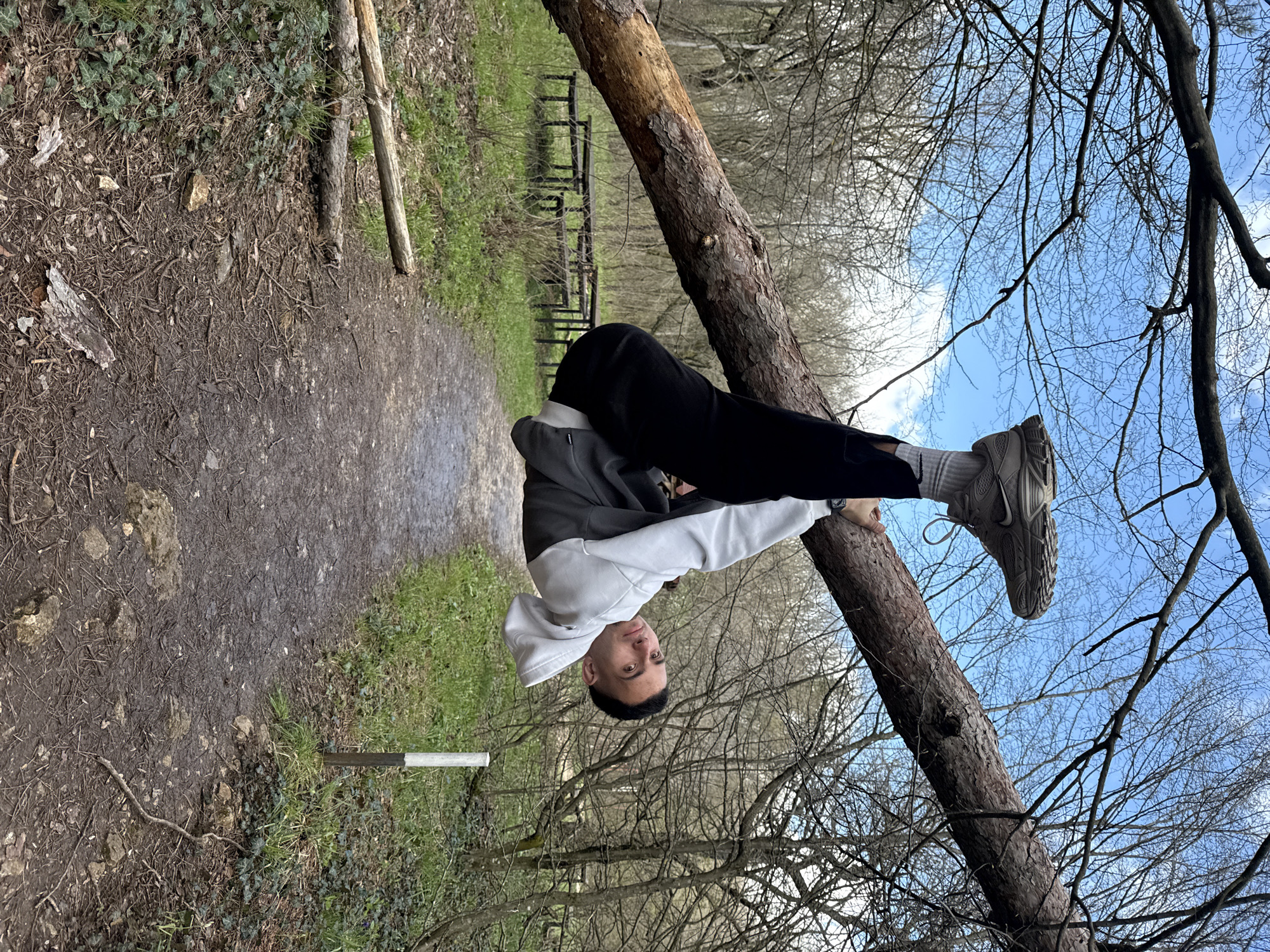 The image depicts a man in a grey and white hooded jacket, black trousers, grey socks, and grey sneakers balancing upside down on a fallen tree trunk in a wooded area. The man is holding onto the tree trunk with his hands and feet, maintaining an inverted position. The background shows a path on the left and a forest with bare trees and a blue sky. The location is in the city of Shumen, Bulgaria.