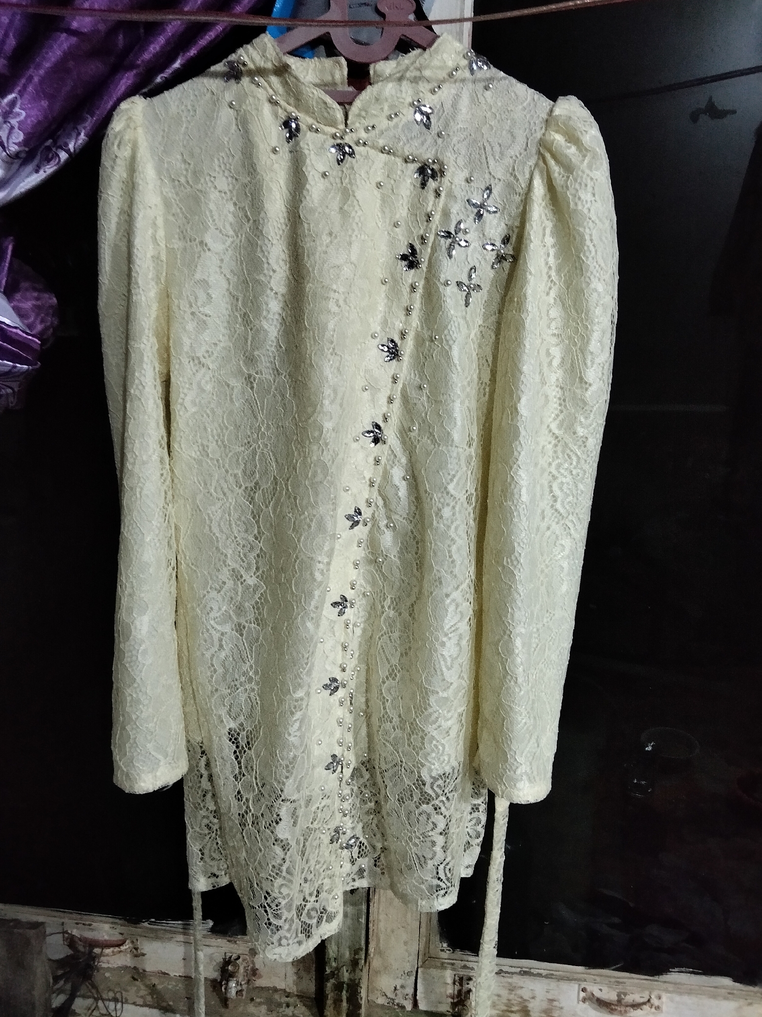 A cream-colored, lace garment is displayed on a hanger. The garment features a high neckline with a mandarin collar and is embellished with small pearls and silver butterfly-shaped appliques. The sleeves are long and puffed at the shoulder. The front of the garment has a diagonal closure adorned with more pearls and appliques. The hemline is uneven and also features pearl and applique embellishments. A cream-colored sash extends from the right side of the garment. The garment is hanging in what appears to be an indoor setting, with a dark background. Part of a purple and white patterned fabric is visible on the left side of the image, and a wooden frame with some debris is visible at the bottom. The image quality is moderate, with some blurriness and low light conditions. There are no people or identifiable landmarks of Benuaraja, Indonesia visible in the image.