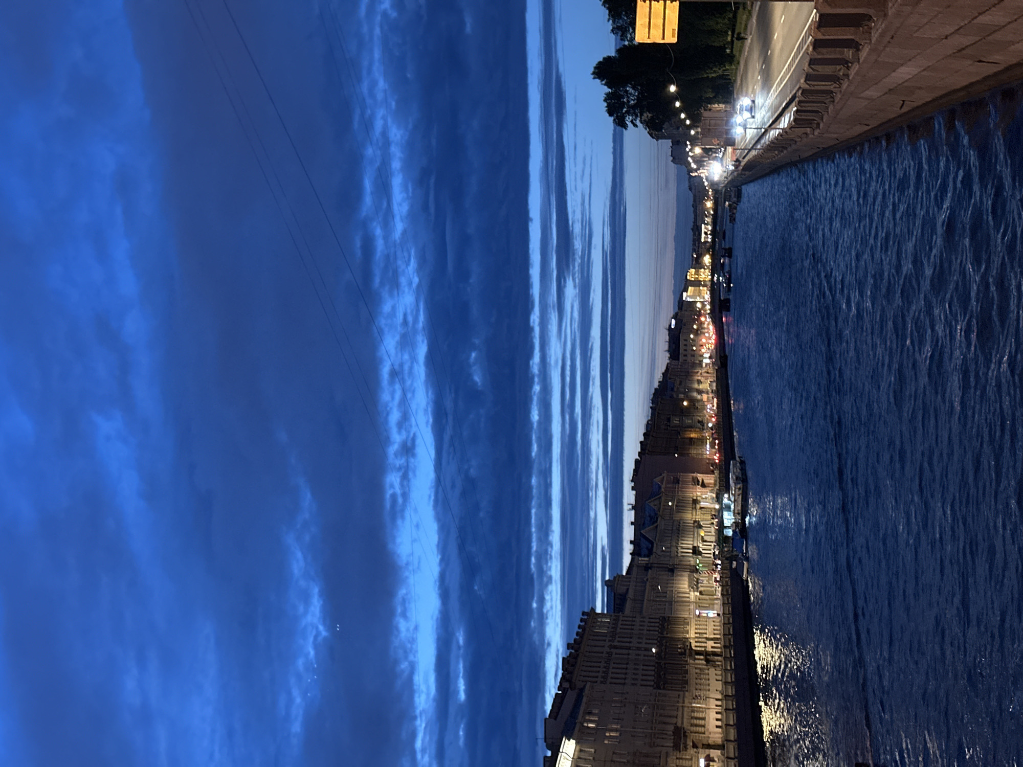 An elevated, wide-angle shot captures a twilight scene along a wide, dark blue river, reflecting the lights of the surrounding city. On the left bank, ornate, multi-story buildings with lit windows and architectural details line the waterfront. Several boats are moored or navigating the river, their lights casting bright reflections on the water. On the right bank, a paved road runs parallel to the river, with streetlights illuminating passing cars. Trees are visible along the embankment, and a yellow sign stands near the road. The sky above is a deep, dark blue, filled with dramatic, layered clouds that create a sense of depth and atmosphere. Two faint pinpricks of light, possibly stars or distant lights, are visible in the upper left quadrant of the sky. The overall impression is one of a vibrant, active city transitioning from day to night.