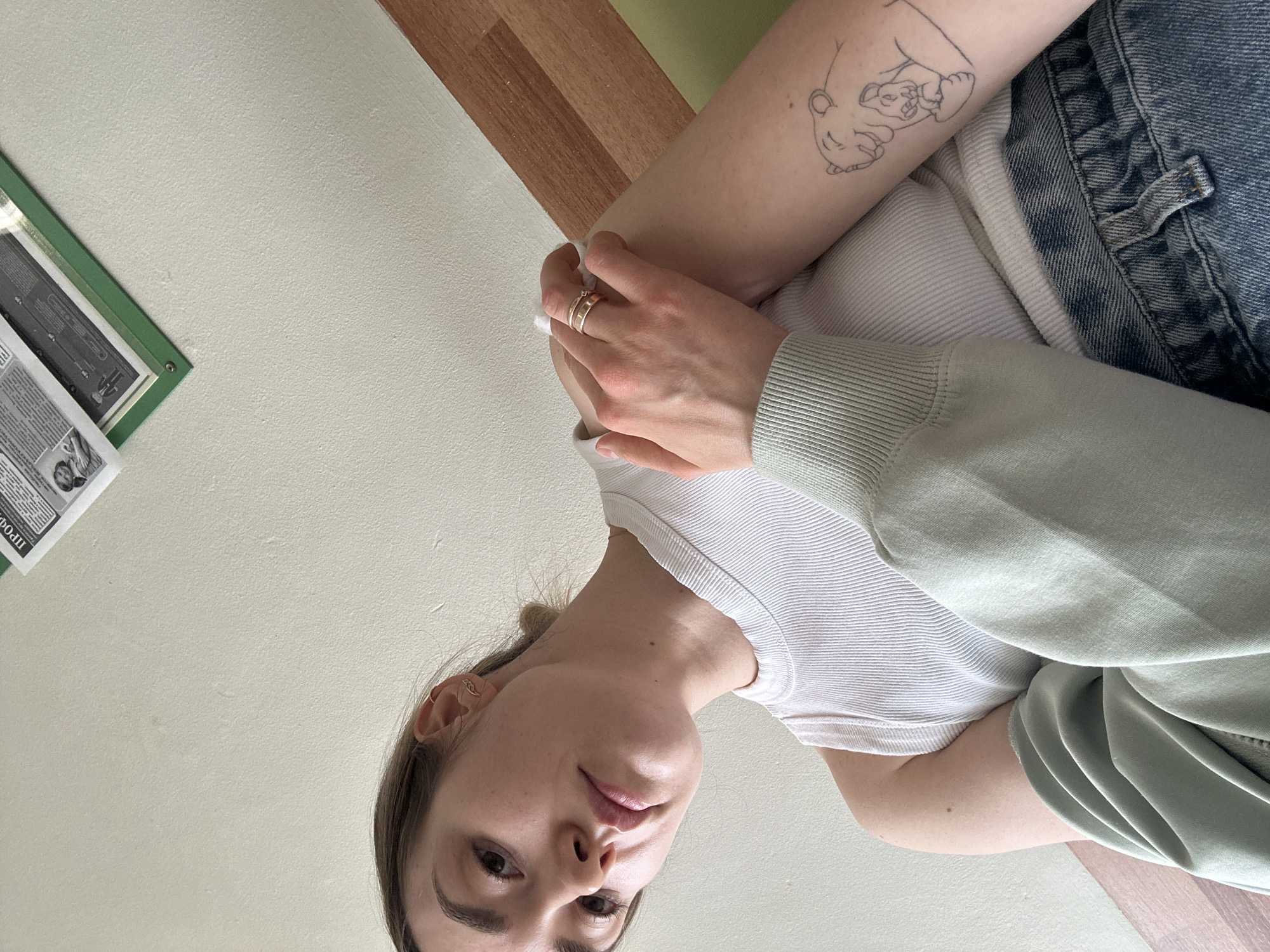 The image shows a woman posing for a selfie indoors. She is wearing a white tank top, a light green sweater, and blue jeans. Her left arm is visible, featuring a tattoo of a sleeping bear. She is wearing rings on her right hand. A framed object is visible on the wall in the upper left corner of the image. The location is not identifiable as Фоки, Russia.