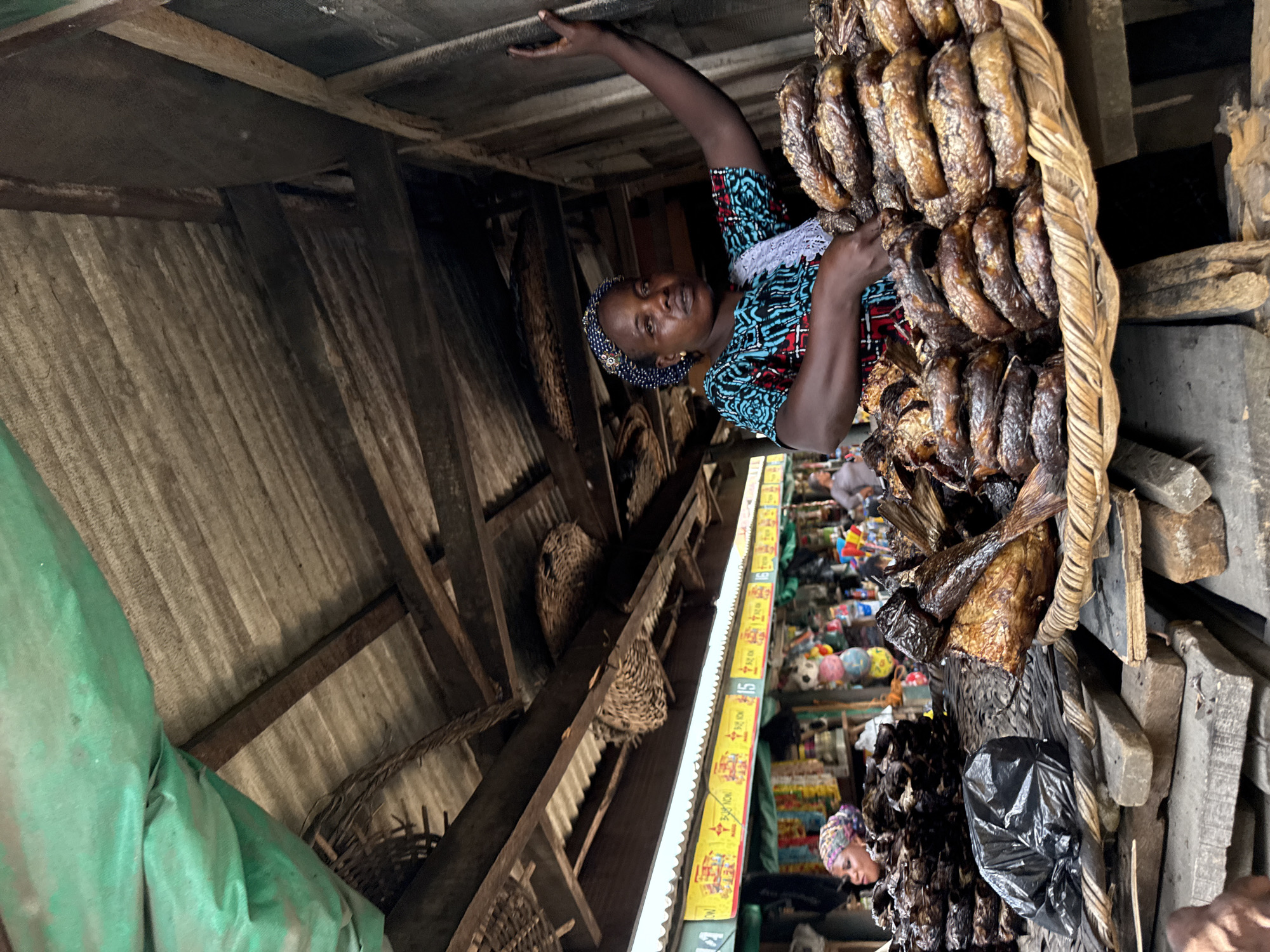 The image captures a market stall in Lekki, Nigeria, focused on the sale of dried or smoked fish. A dark-skinned female vendor, positioned centrally, wears a blue patterned headscarf and a geometric-patterned teal, red, white, and black top with white lace trim. She looks directly at the viewer. Her right arm is raised, with her hand resting on a wooden wall or partition to the right. Her left hand is positioned over large quantities of dark-brown, dried fish, which are neatly stacked in circular piles on large, round, woven trays. These trays are supported by a structure of stacked rough wooden planks. To the left and slightly behind her, another dark-skinned woman with a colorful patterned headscarf is partially visible, facing left, behind a separate pile of similar fish. A black plastic bag is present on the wooden support structure. The stall features an overhead structure composed of corrugated metal sheets supported by wooden beams, with a green tarp partially visible in the upper left corner. The background reveals a bustling market scene with other stalls displaying various goods, indistinct people, and yellow banners with text including "BUY NOW" and numbers "15", "16".