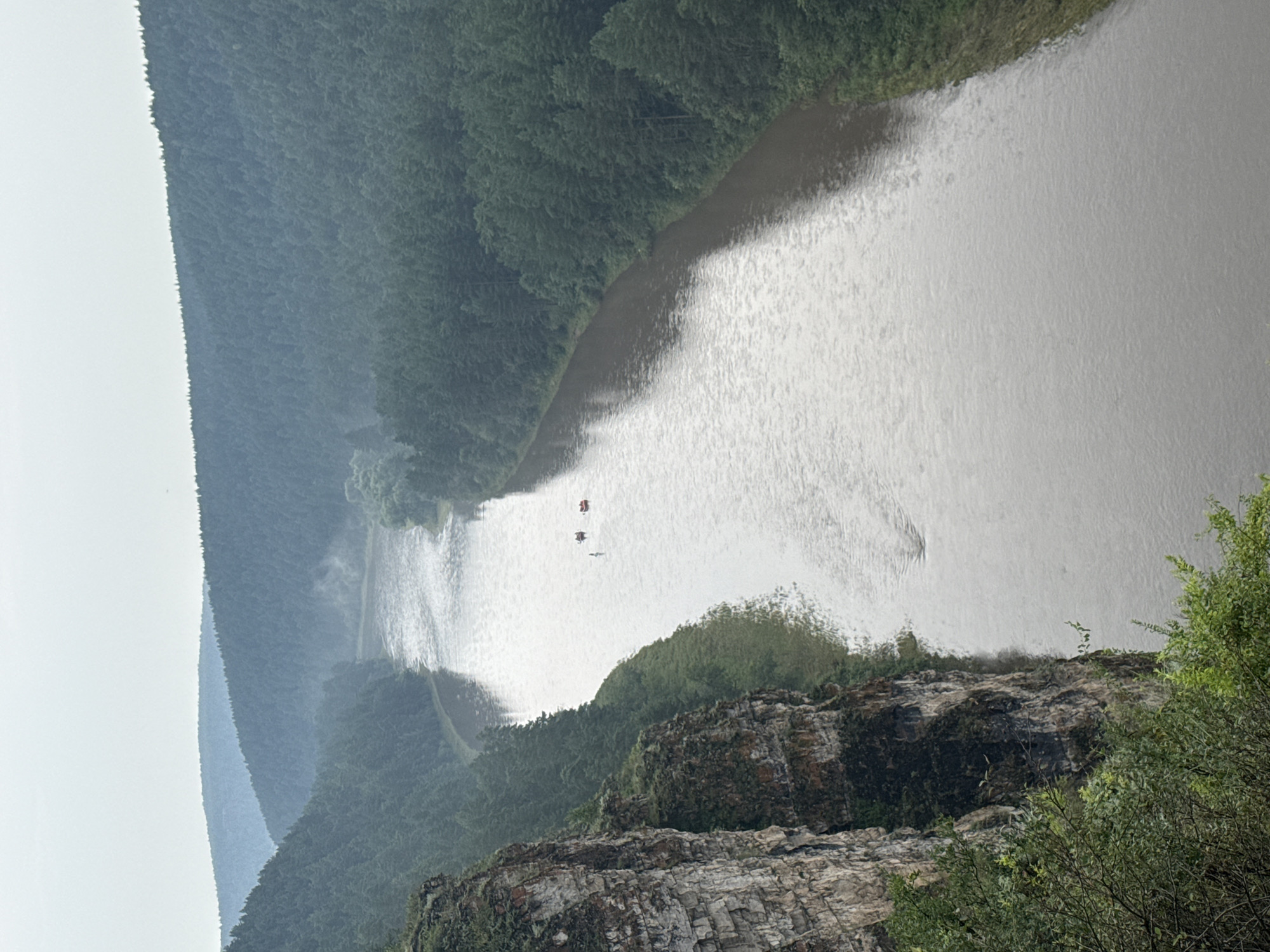 This is a high-angle, medium shot of a wide, muddy river flowing through a forested valley on an overcast day. A steep, rocky cliff face with some sparse vegetation lines the left side of the frame, dropping down to the river's edge. The right side of the river is bordered by a dense forest of dark green coniferous trees that stretch up the hillsides. In the distance, the hills recede and become shrouded in mist, with multiple layers of blue-hued, forested mountains visible. On the river, approximately mid-frame, are three small, orange-red inflatable rafts or boats, each with at least one person visible, suggesting recreational activity. The water is choppy with ripples and some surface debris is visible. The overall atmosphere is cool and damp. The location is identified as the city of Kul'metovo, Russia.