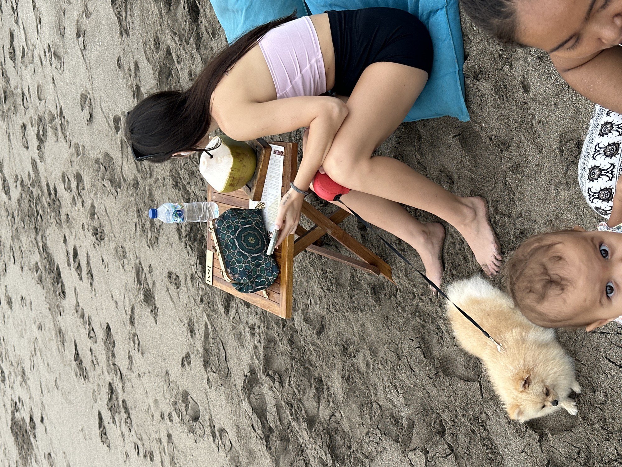 A young woman with long dark hair is sitting on a blue cushion on a sandy beach, likely in North Kuta, Indonesia, as indicated by the presence of a young child and a small dog on a leash. The woman is wearing a light pink tube top and black shorts and is holding a smartphone. In front of her is a small wooden table with a patterned clutch, a bottle of water, and a halved coconut. A small, fluffy beige dog is lying on the sand next to her, attached to a black leash. A baby is also visible in the foreground, looking towards the camera. The beach is covered in sand with visible footprints.