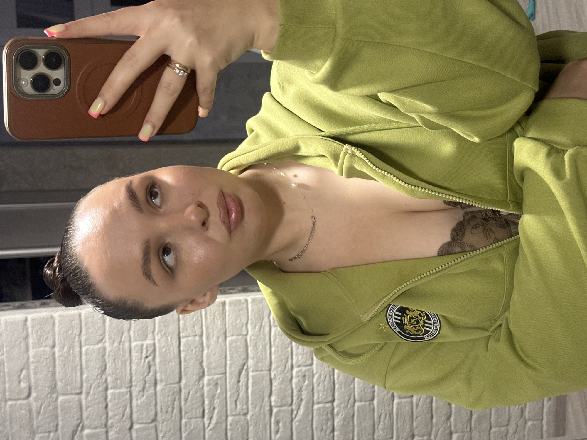 A woman is taking a selfie in what appears to be an indoor setting. She is holding a smartphone in her left hand, with the camera facing her. She has her hair pulled back in a bun and is wearing a light green zip-up jacket, partially unzipped, revealing a lace garment underneath. She has a necklace on. The background shows a white brick wall. There is no specific evidence to indicate she is in Chaikovsky, Russia.