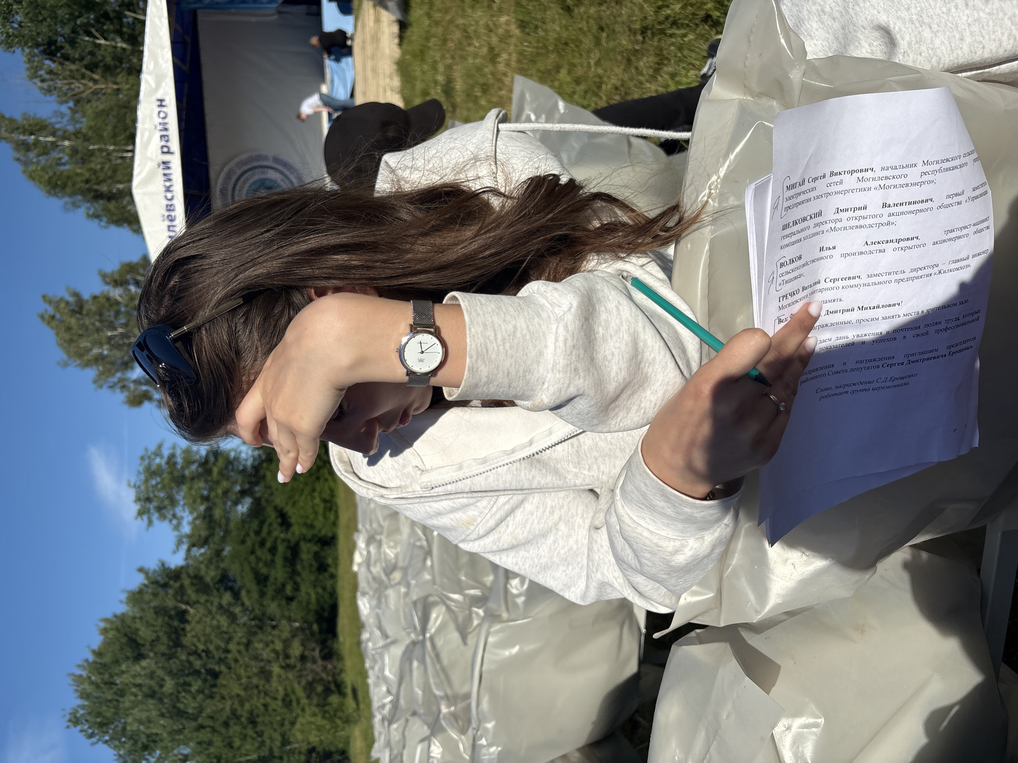 This is a vertical, medium shot of a young woman sitting outdoors, looking down at a piece of paper in her hand. She has long, dark hair that falls over her face, obscuring her features. She is wearing a light-colored hooded sweatshirt and a watch on her left wrist. Her right hand holds a green pencil, and she appears to be writing or reviewing the document. The paper she is holding has text in Russian, identifying individuals and their positions within various organizations, likely related to local administration or public service.
The setting appears to be a sunny outdoor event, with a blue sky and green trees visible in the background. A white tent or banner with text in Russian is also partially visible, suggesting a public gathering or ceremony. The woman is seated on what appears to be a white, textured surface, possibly part of a seating arrangement or stage setup. The overall impression is one of a formal event taking place on a pleasant day. The text on the paper suggests a recognition or award ceremony.