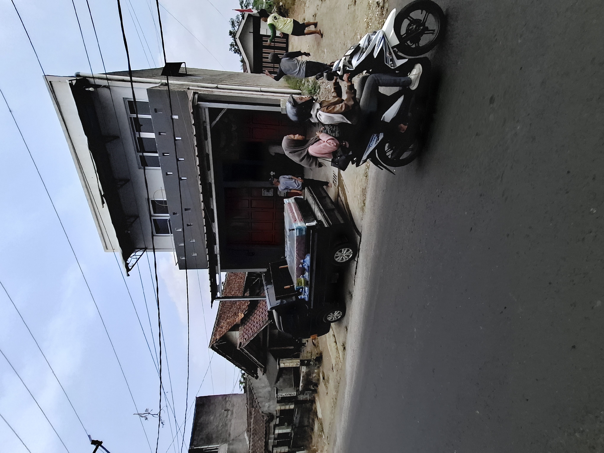 The image depicts a street scene in Nagrak, Indonesia. In the foreground, a white and black scooter is being ridden by two individuals, a driver wearing a helmet and a passenger behind them. They are moving along a paved road. To the left of the road, a black pickup truck is parked, loaded with what appears to be mattresses and several blue water jugs. Behind the truck, a modest building with a red tiled roof is visible. Further back, a multi-story building with a modern design and dark-colored awnings stands. The sky is overcast. Several people are present near the buildings, some walking and others standing. One individual is wearing a yellow shirt and appears to be interacting with another person. Power lines are strung across the upper portion of the image.