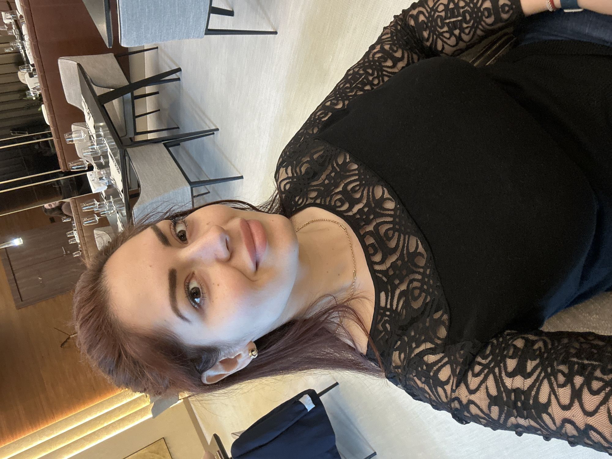 A medium shot depicts a woman with fair skin, dark hair, and brown eyes, situated indoors. She is wearing a black top with lace sleeves, as well as a gold necklace. She is photographed from a slight angle, presenting a partial view of her face.
The background reveals a restaurant setting, with tables and chairs arranged neatly. A close inspection suggests this location might be in Kazanlak, Bulgaria, though this is not definitively confirmed. The lighting appears to be a mix of natural and artificial sources, creating a moderately illuminated scene.