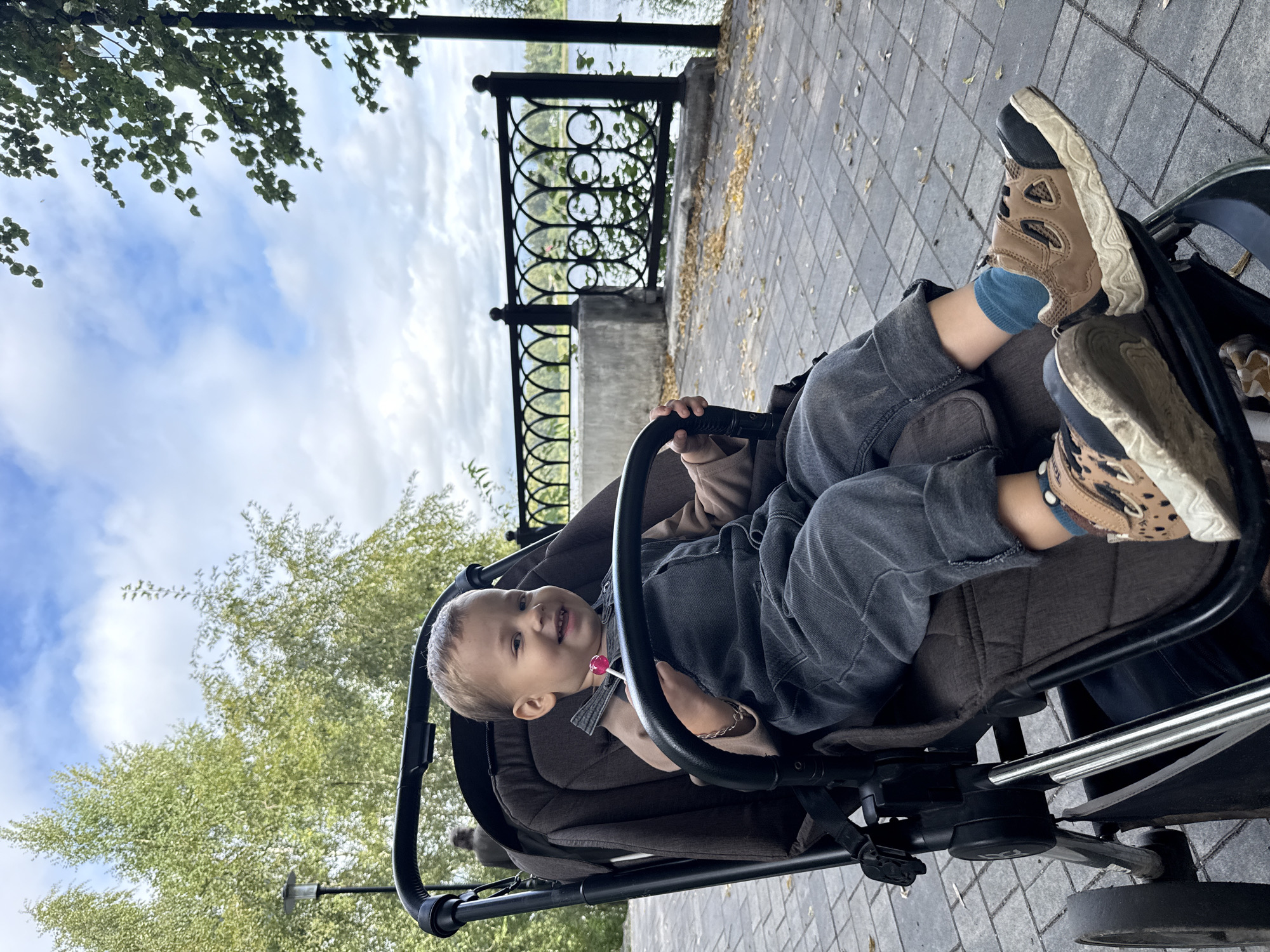 A young child is seated in a stroller on a paved pathway next to a body of water. The child, a boy with light brown hair, is wearing a beige long-sleeved shirt, dark grey overalls, blue socks, and brown and black sneakers with a leopard print detail. He is smiling and holding a pink lollipop. The stroller is dark brown with a black frame and wheels. In the background, there is a black metal fence, trees, and a cloudy sky. A person can be seen in the distance behind the child. The setting appears to be a park or promenade in the city of Dumino, Russia.