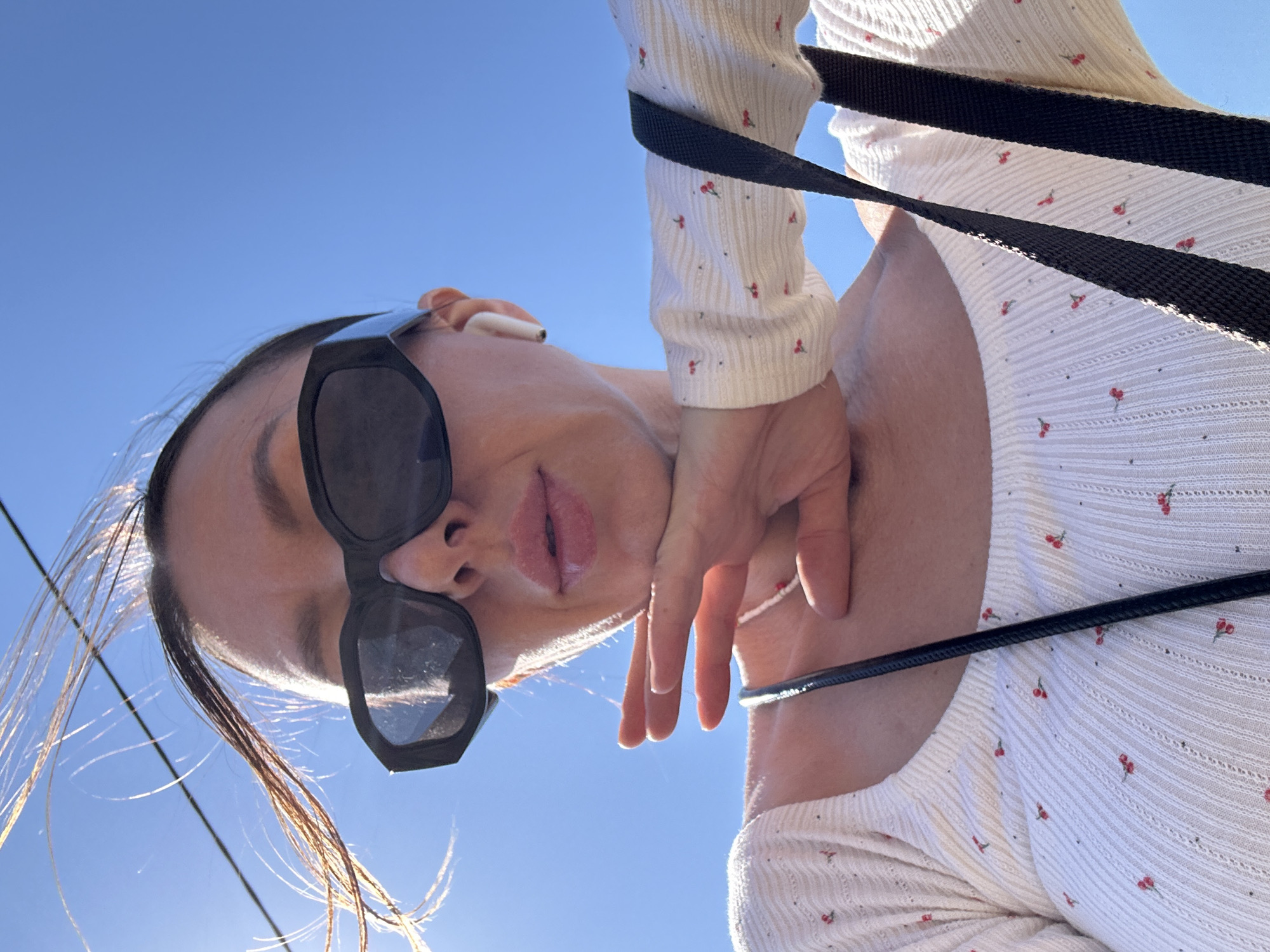 A person is angled upward against a clear blue sky. The person is wearing sunglasses and an earbud is visible in their ear. They are wearing a white, long-sleeved shirt with small cherry designs. A black strap is draped across their body. The location is not discernible from the image.